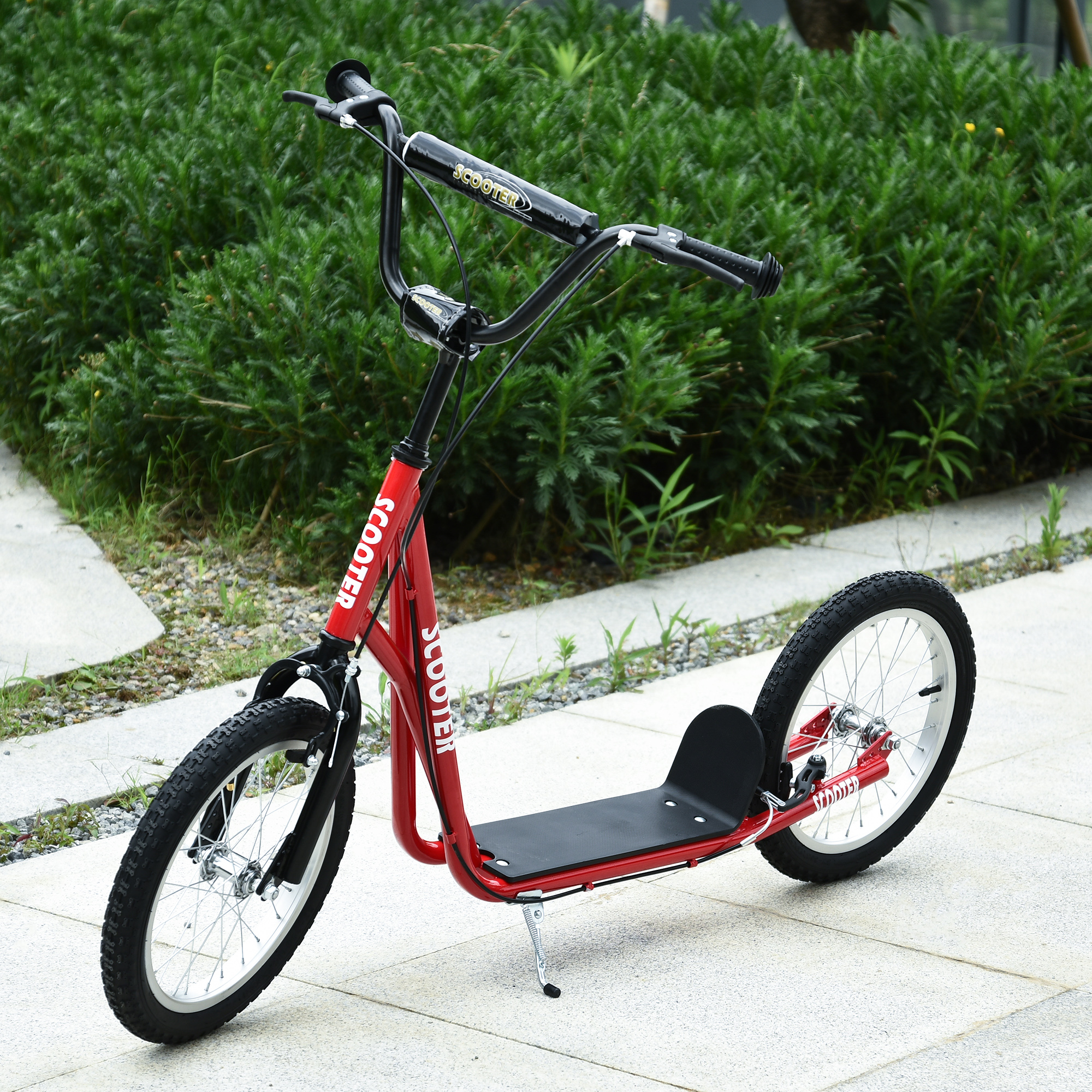 Un patinete rojo con manillar negro sobre pavimento, arbustos verdes al fondo.