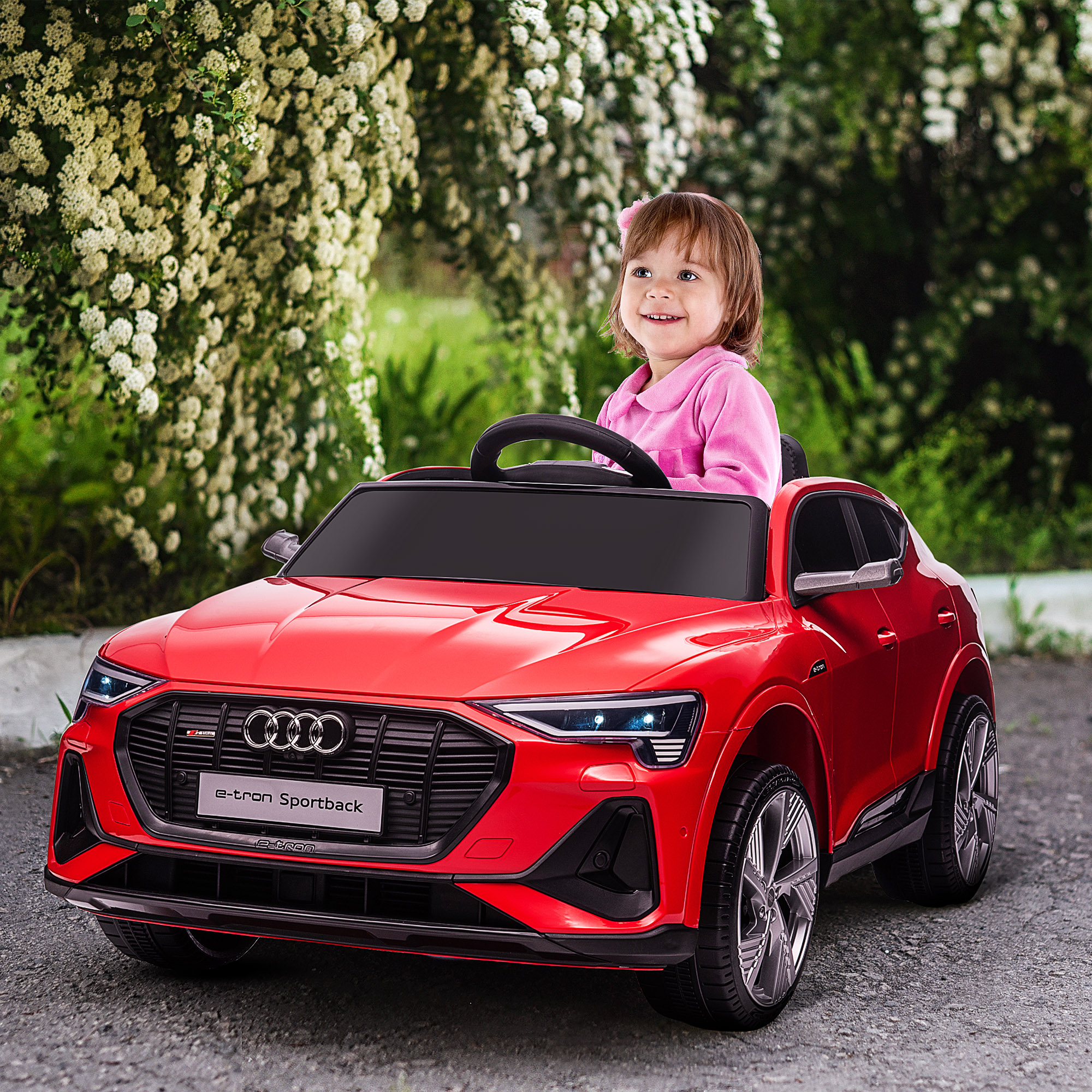 Bambina su un'Audi giocattolo rossa sorride mentre guida. Fogliame verde sullo sfondo.