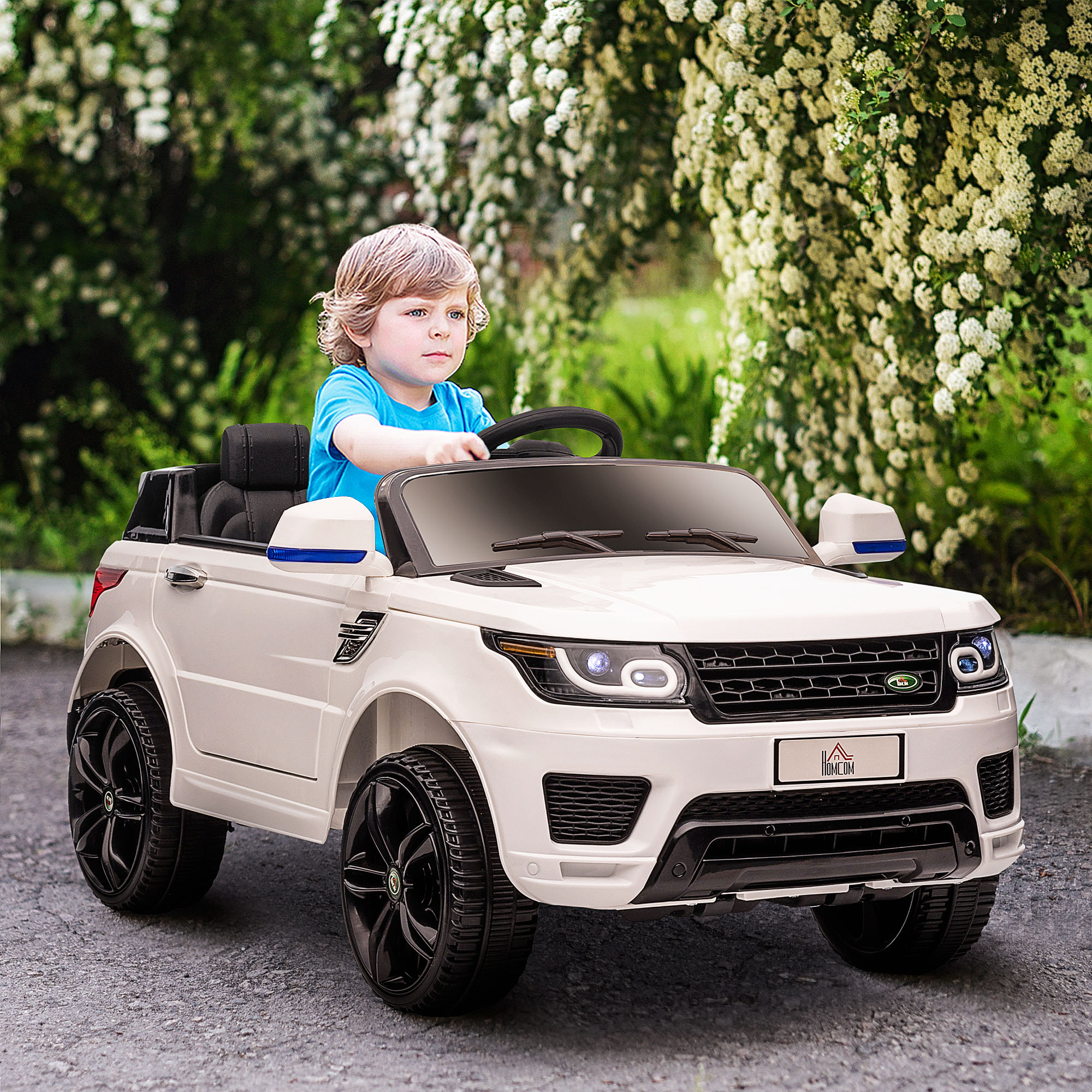 Un bambino guida un'auto giocattolo bianca su una strada asfaltata, con vegetazione sullo sfondo.