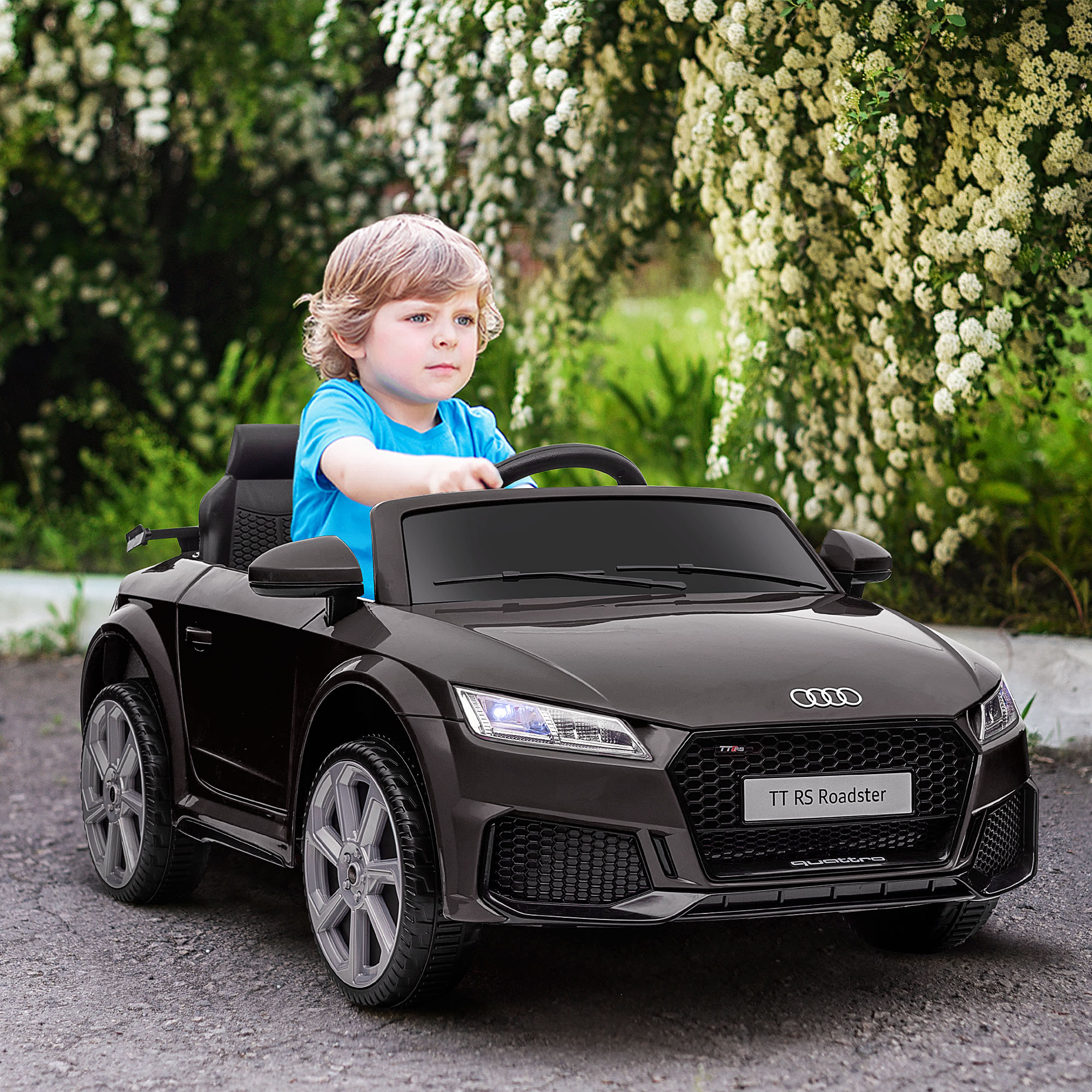 Un niño pequeño conduciendo un coche negro. El coche es un modelo TT RS Roadster. Está en una carretera pavimentada.