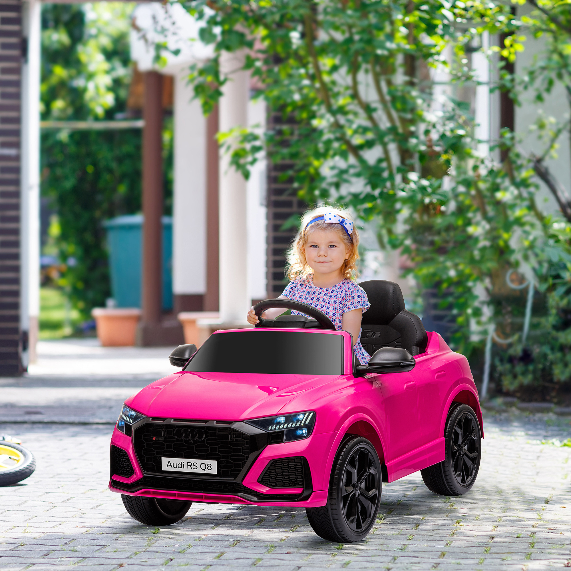 Una niña en un coche de juguete Audi RS Q8 rosa. Árboles verdes y casas al fondo.