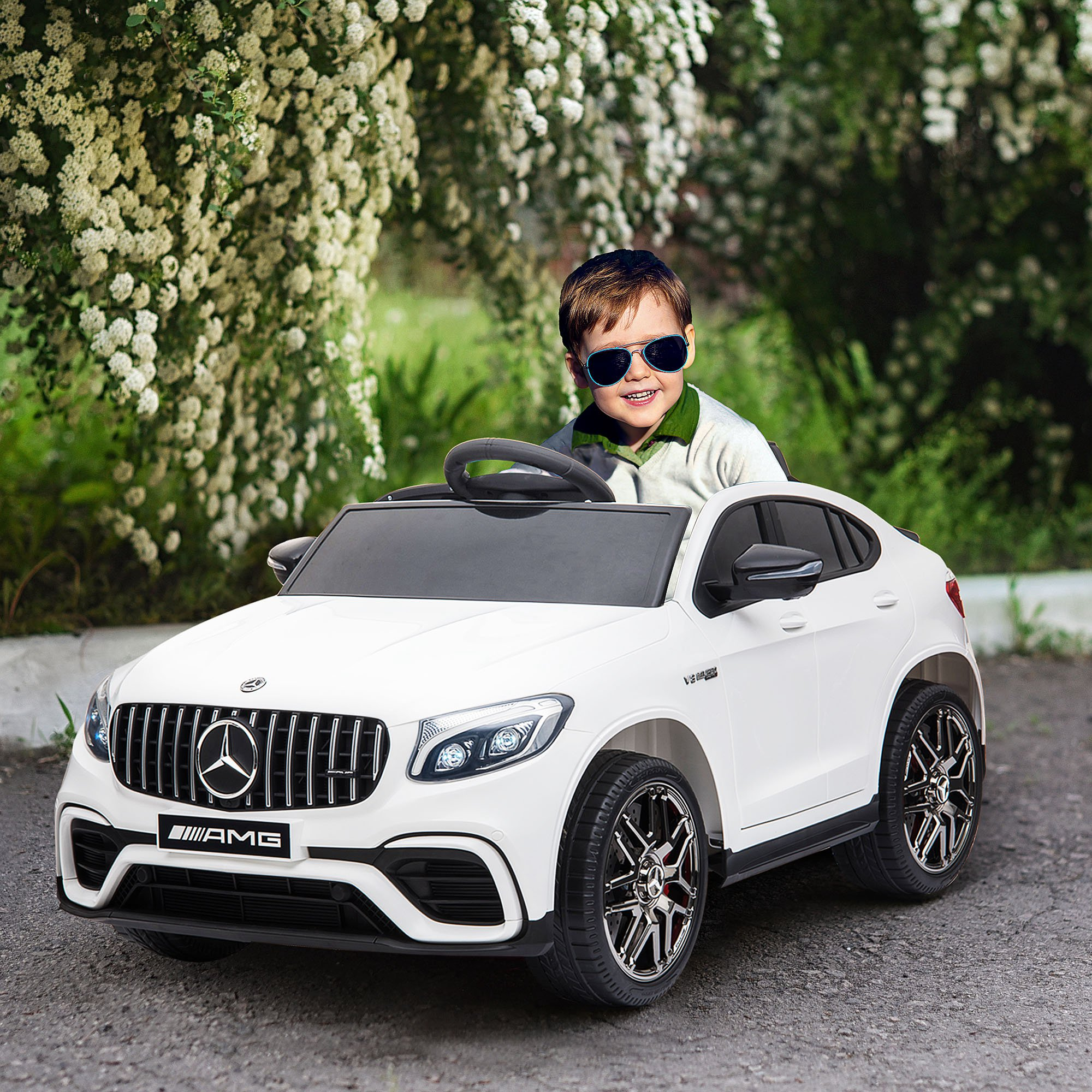 Un niño pequeño con gafas de sol está sentado en un coche de juguete blanco con detalles negros, en un entorno verde.