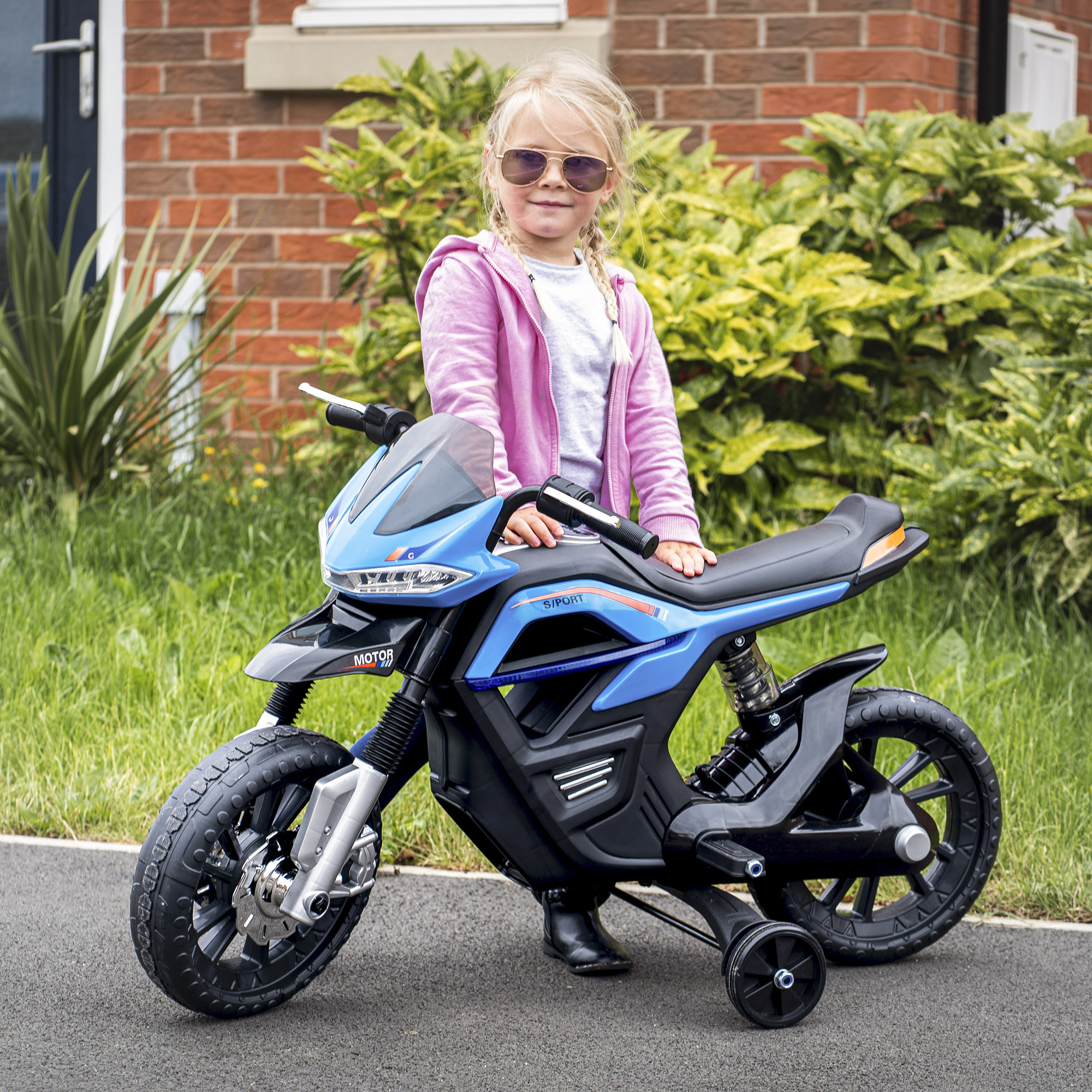 Ragazza con occhiali da sole su una moto giocattolo blu, fuori da una casa in mattoni.