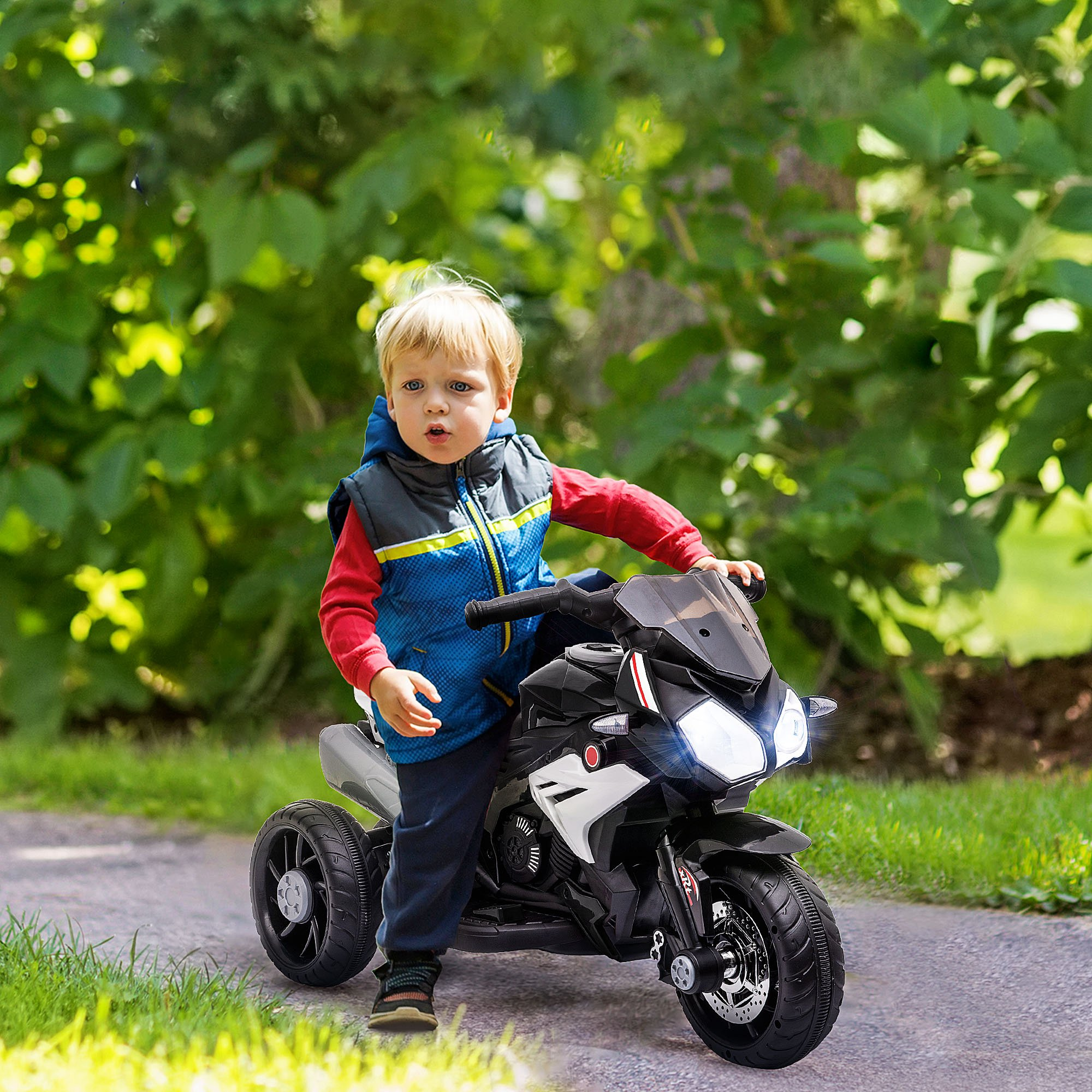 Niño junto a una moto de juguete negra. Follaje verde al fondo. Luces encendidas. Niño con chaleco azul.