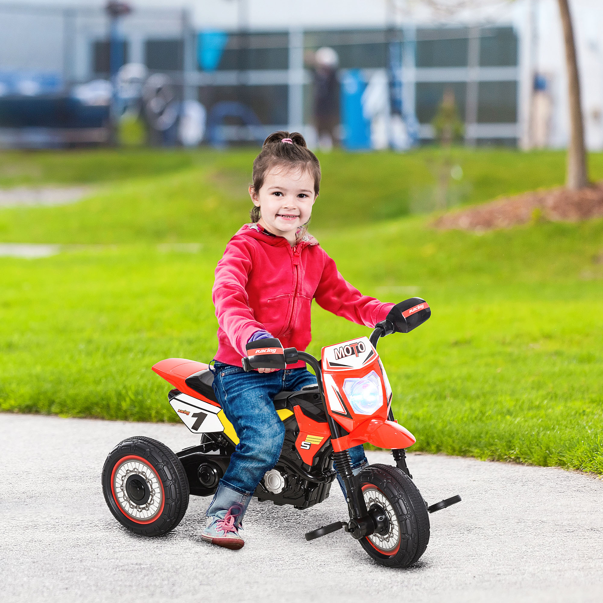 Bambina che guida una moto giocattolo rossa. La ragazza indossa giacca rossa e jeans. Sfondo di erba verde.