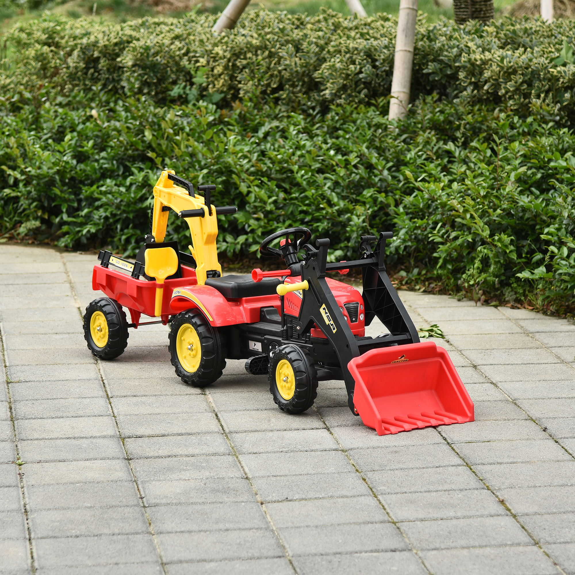 Un tractor de juguete amarillo y rojo sobre una superficie pavimentada, con un remolque rojo. Arbustos verdes al fondo.