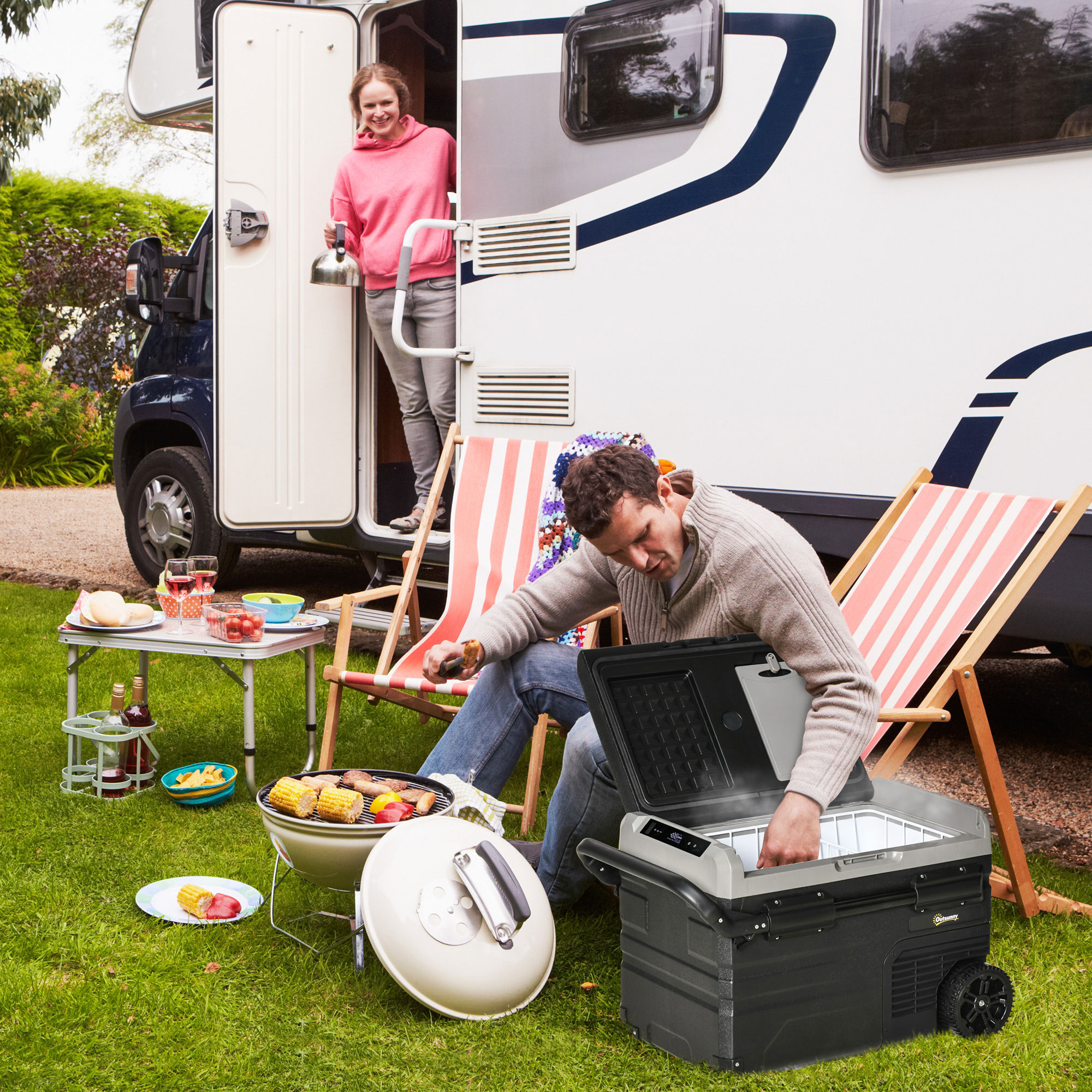 Un hombre asando a la parrilla y una mujer en una caravana. Comida, bebidas y sillas de camping son visibles.