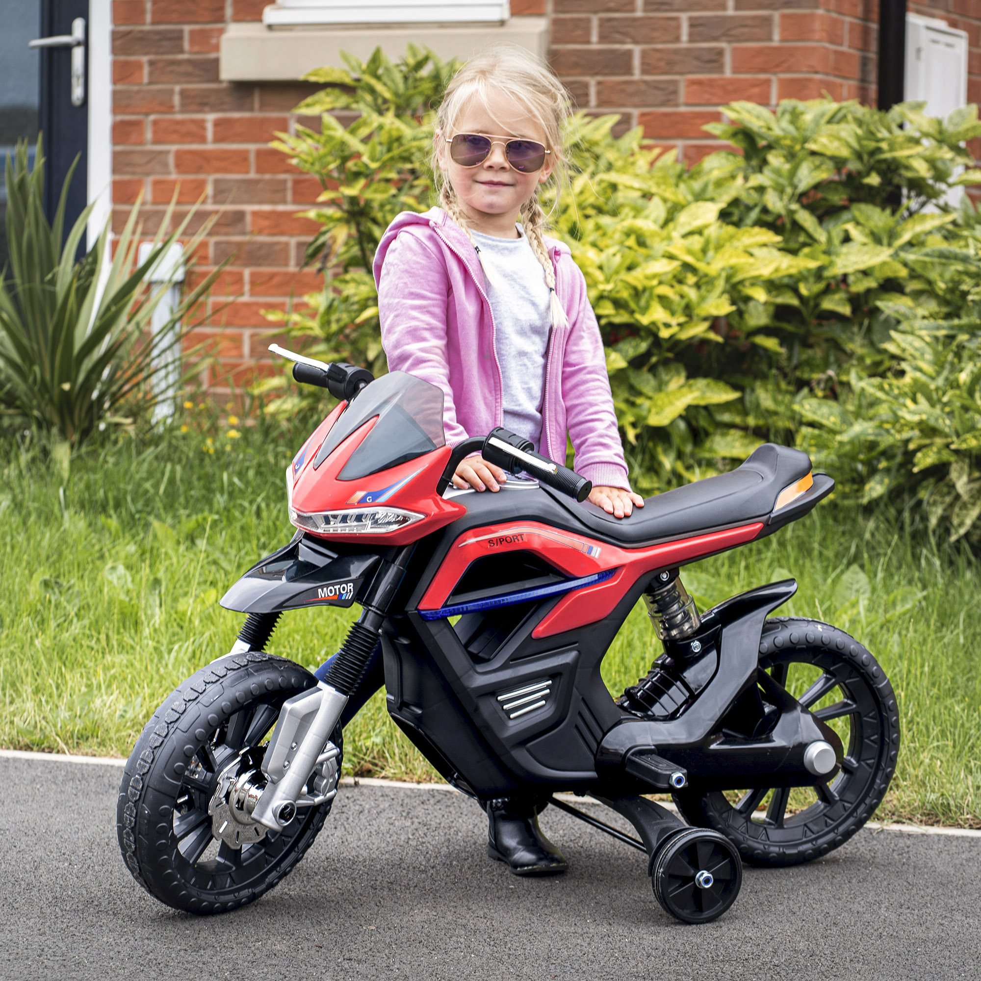 Una niña con gafas de sol está junto a una moto de juguete roja y negra.