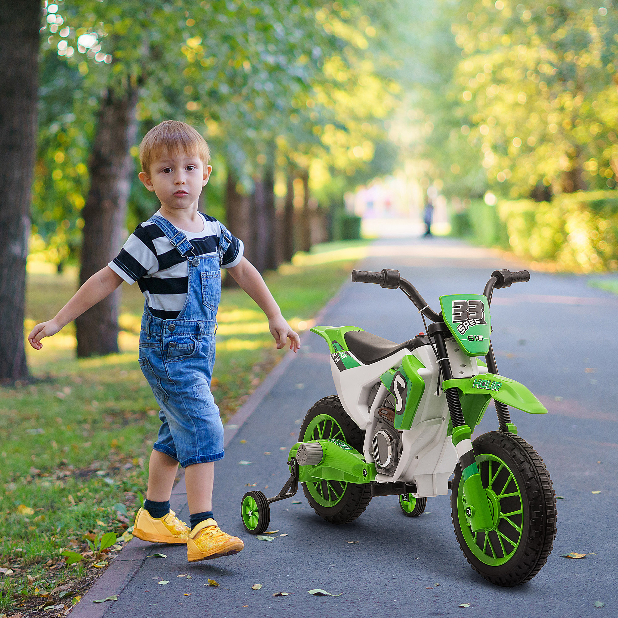Un ragazzino con una moto giocattolo. È in un parco, vicino a una moto verde e bianca con ruote di supporto.
