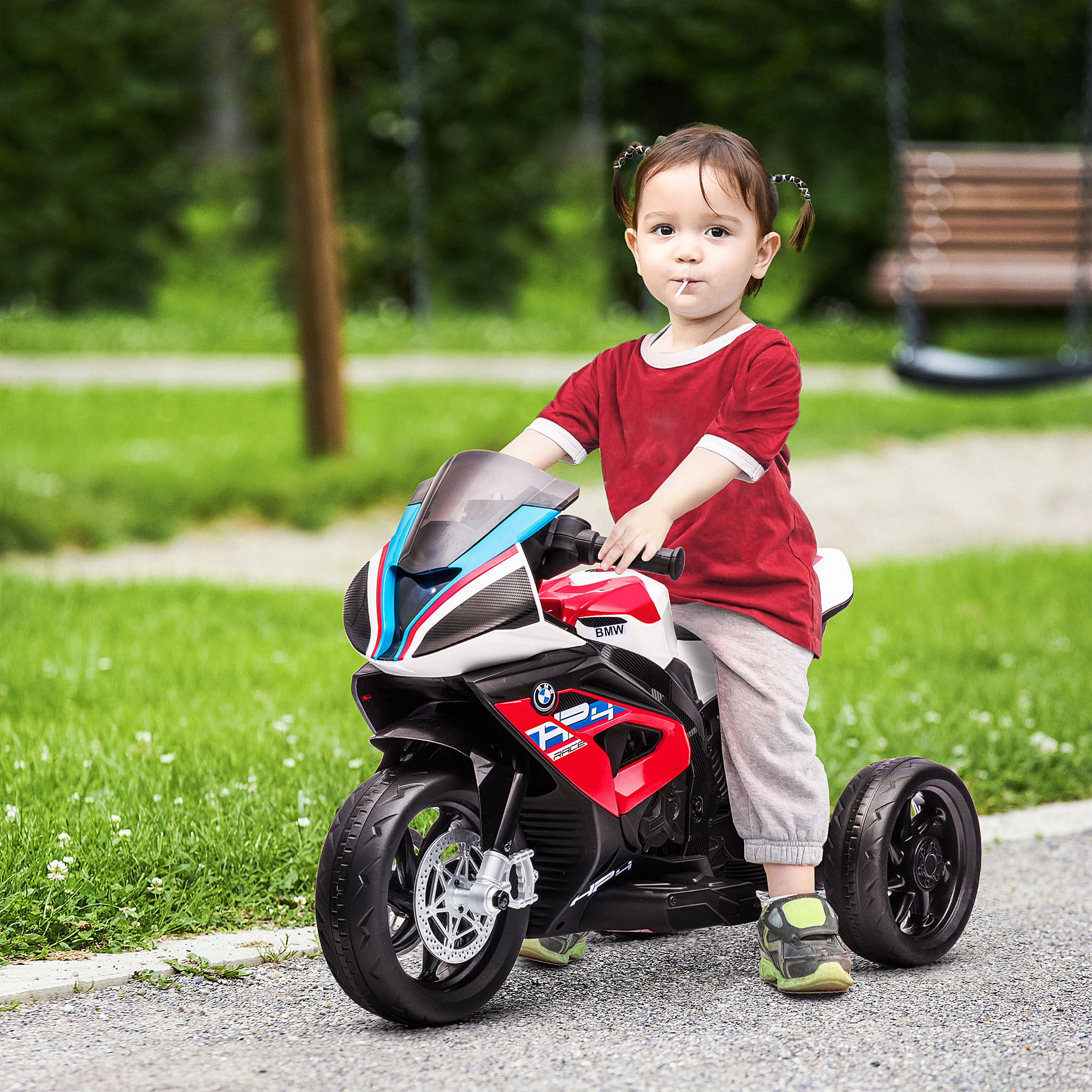 Una niña pequeña anda en una moto de juguete roja, blanca y azul sobre un camino pavimentado. Lleva una camisa roja.