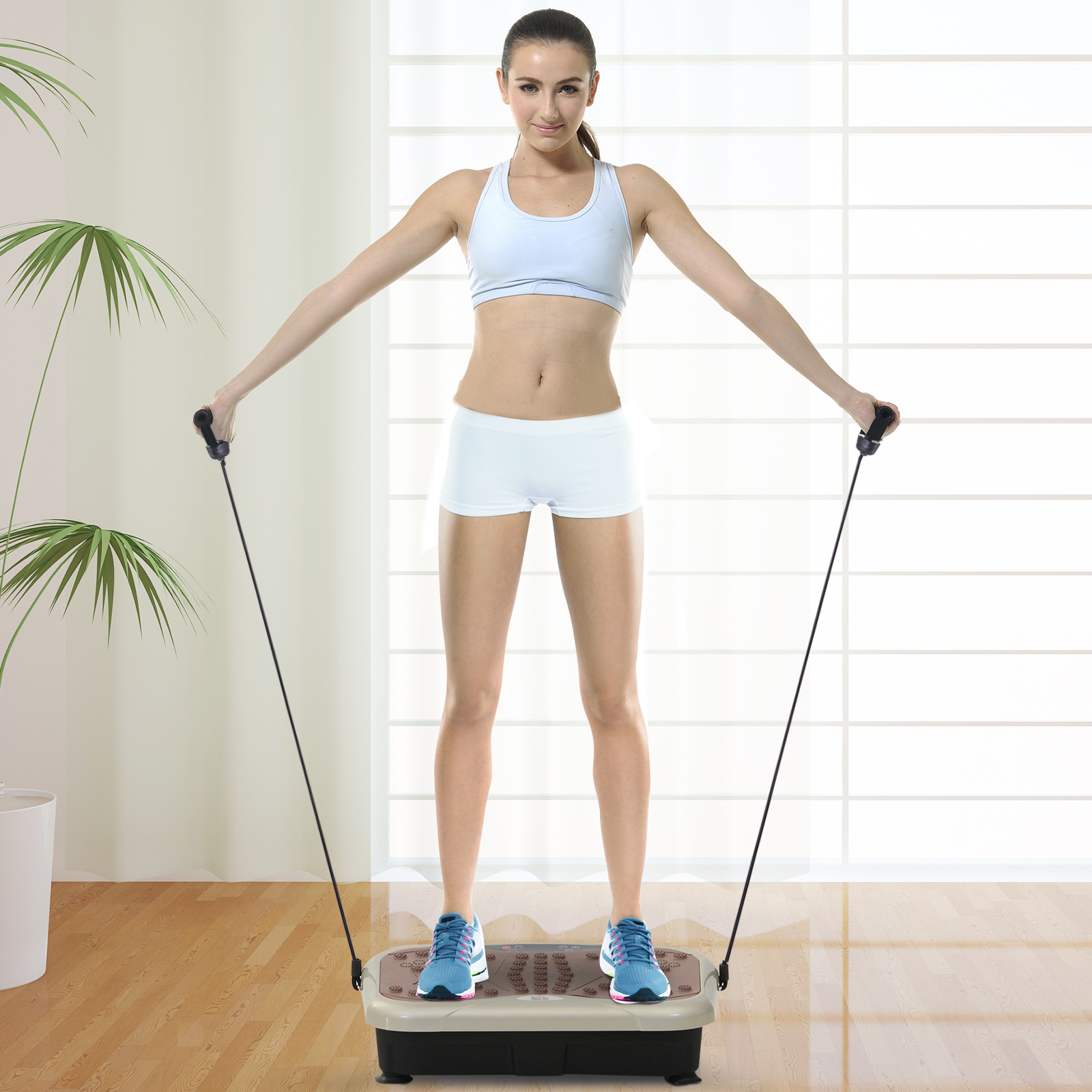 Una mujer con sujetador y shorts deportivos blancos está de pie sobre una plataforma vibratoria, sujetando bandas elásticas.