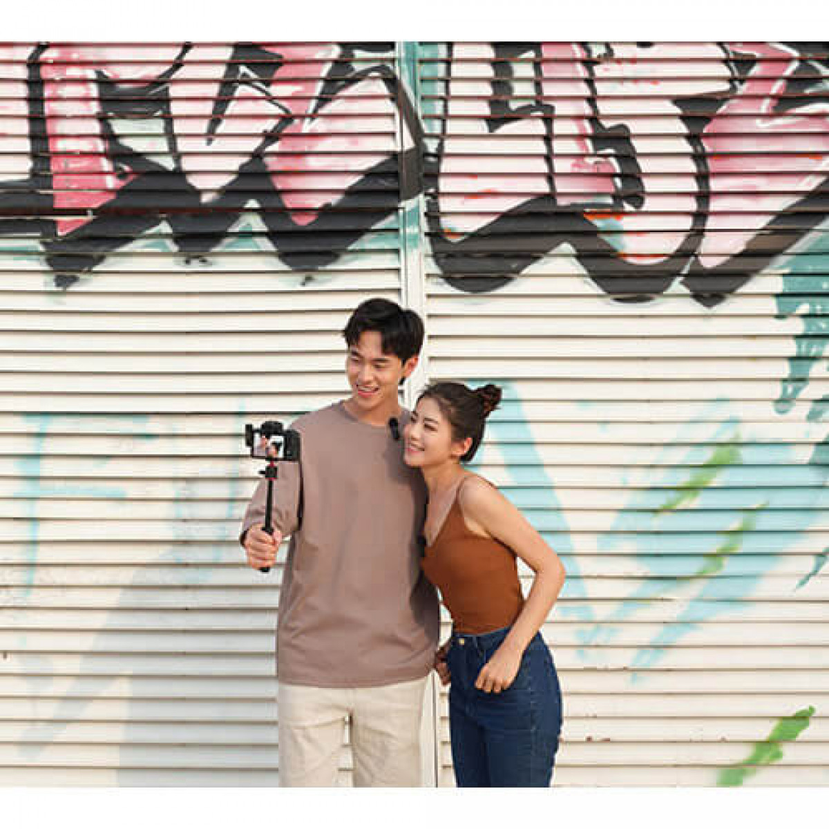 Una coppia posa con una fotocamera su un supporto. Sono di fronte a dei graffiti. L'uomo tiene la fotocamera.