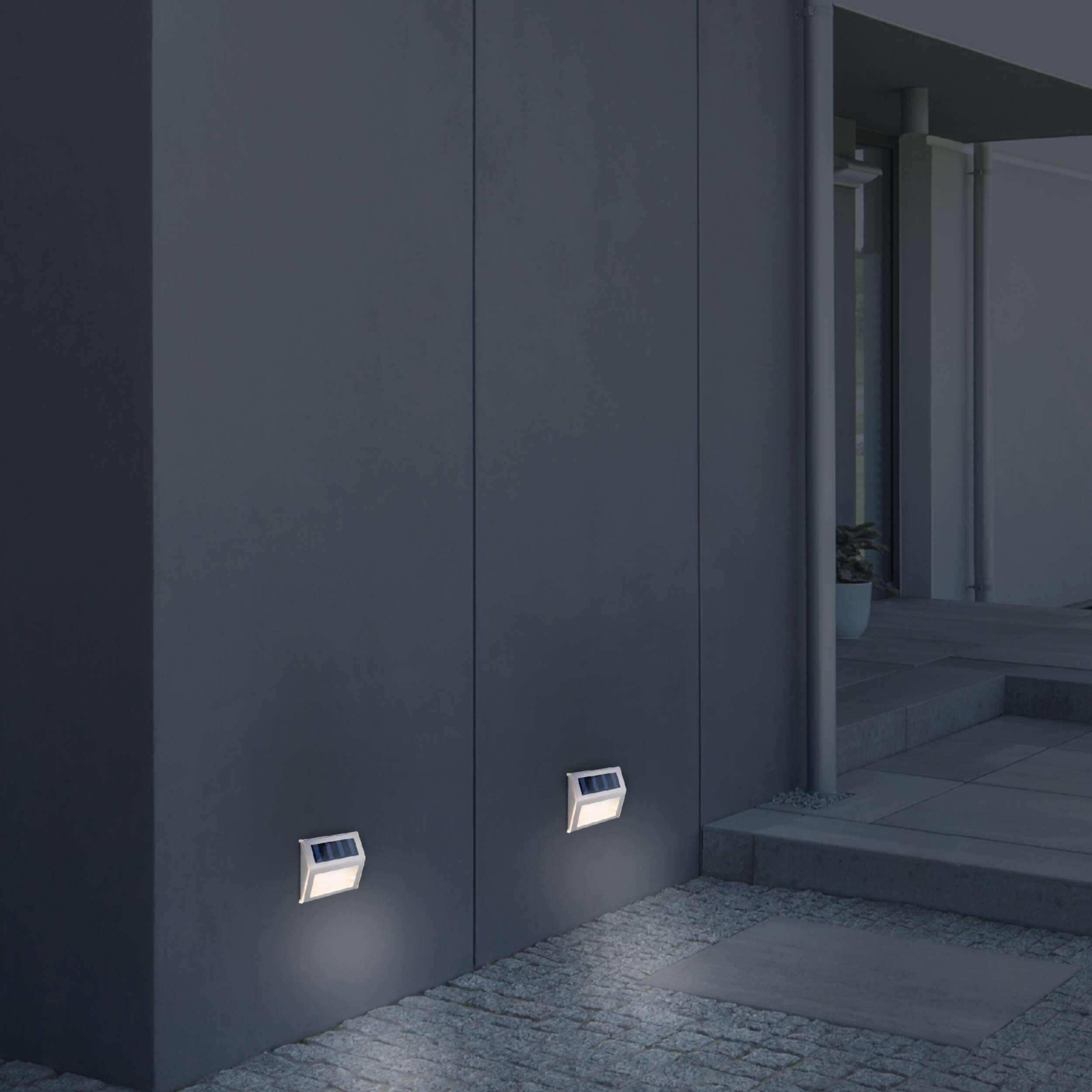 Zwei wandmontierte Solar-LED-Leuchten, die leuchten. Eine dunkelgraue Wand und ein Steinweg im Hintergrund.