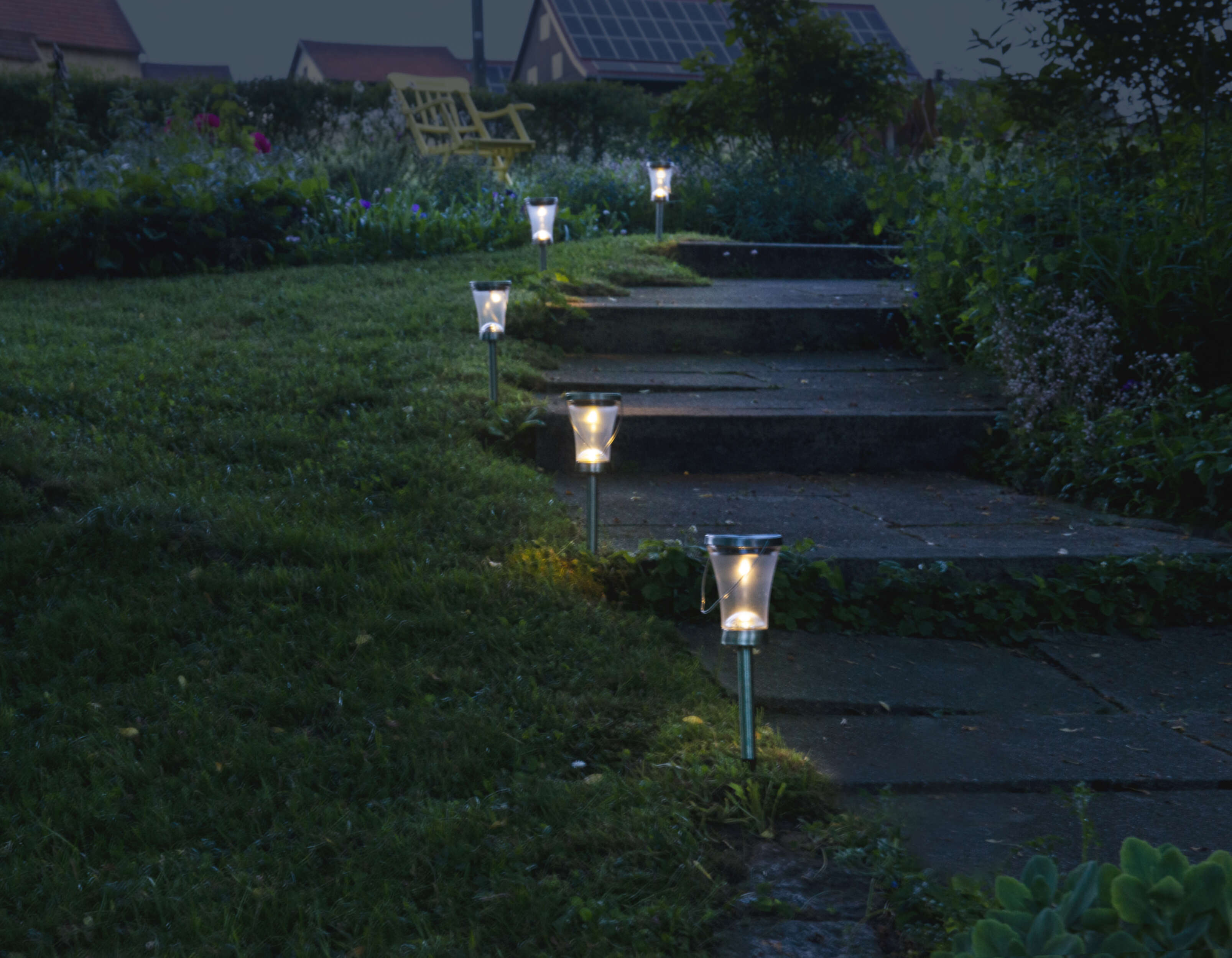 Weg mit Gartenlichtern in der Dämmerung. Gras, Stufen und Lichter erzeugen eine einladende Szene.