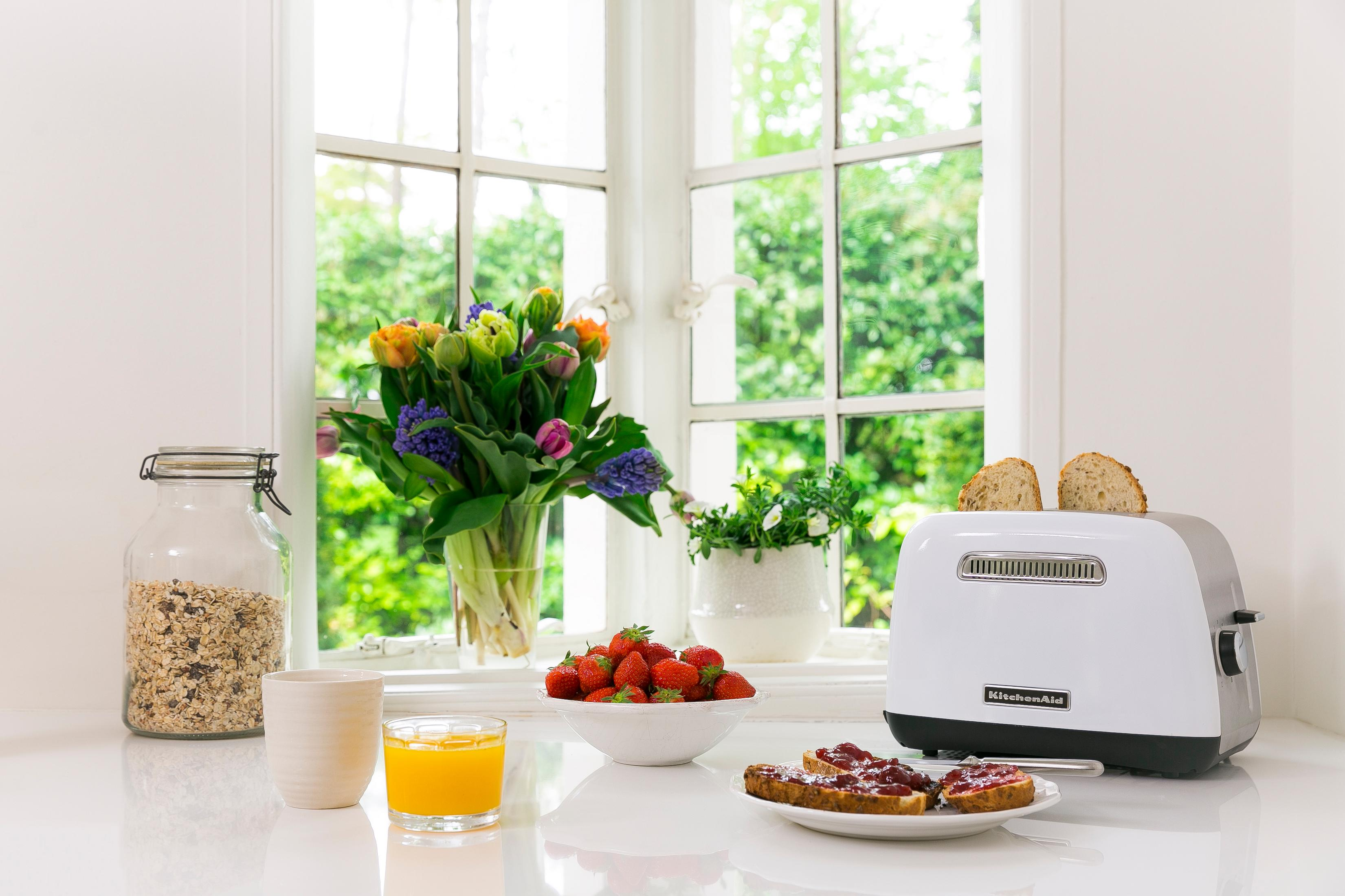 Kitchen counter with a toaster, flowers, fruit, and a jar. Window in background.