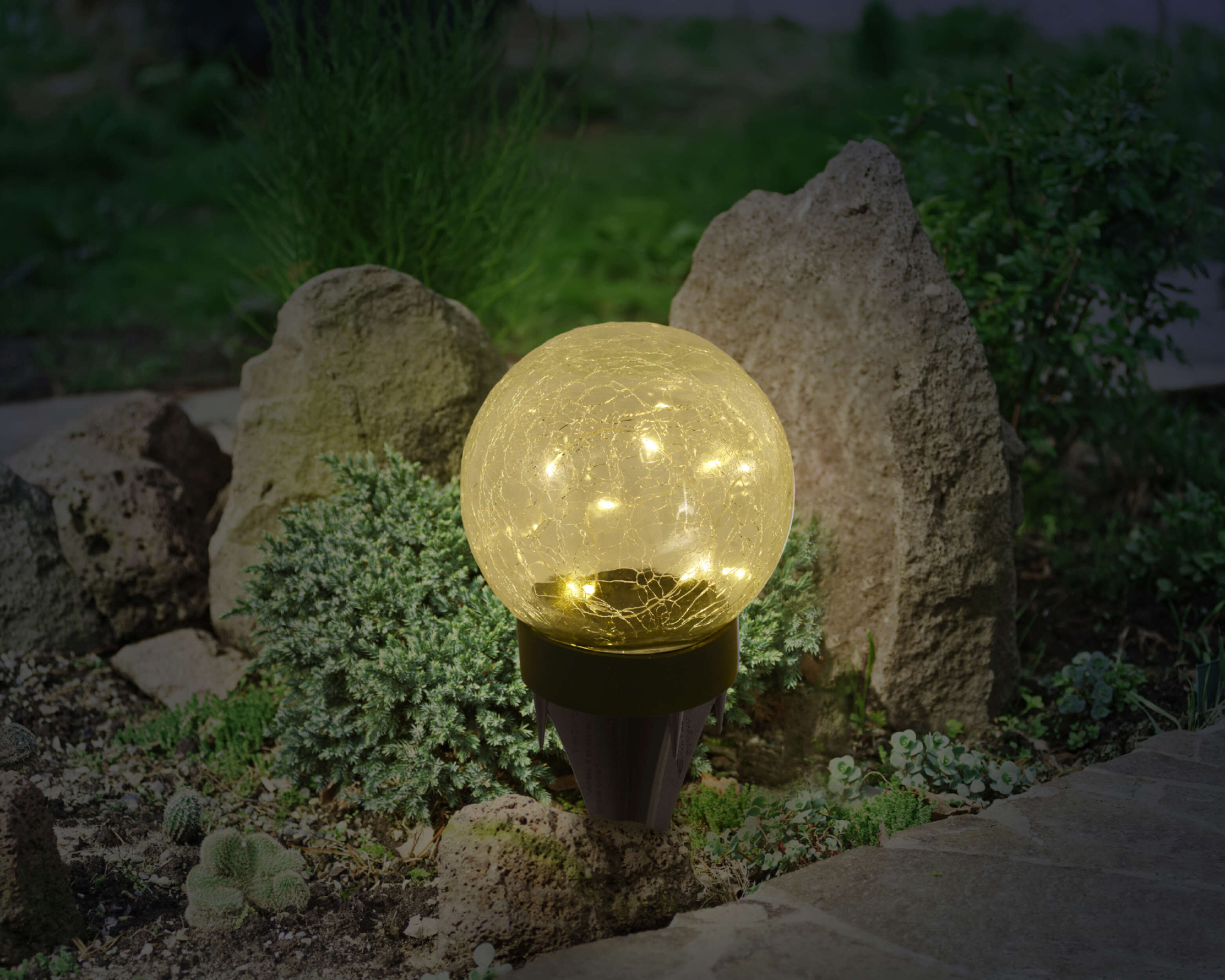 Solarbetriebene Gartenleuchte, eine beleuchtete Glaskugel mit Pflanzen im Hintergrund.
