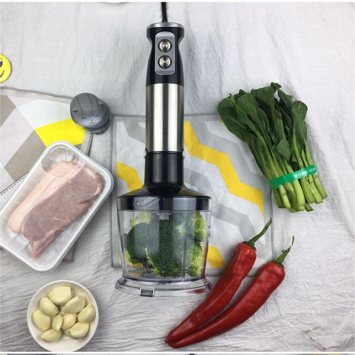 Handmixer mit Brokkoli, roten Paprika, Schweinefleisch, Knoblauch und Grünzeug auf weißer Oberfläche.