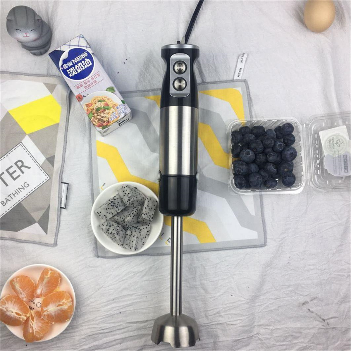 Eine Lebensmittelszene mit einem Mixer, Früchten und einem Sahnekarton auf einem weißen Tuch.
