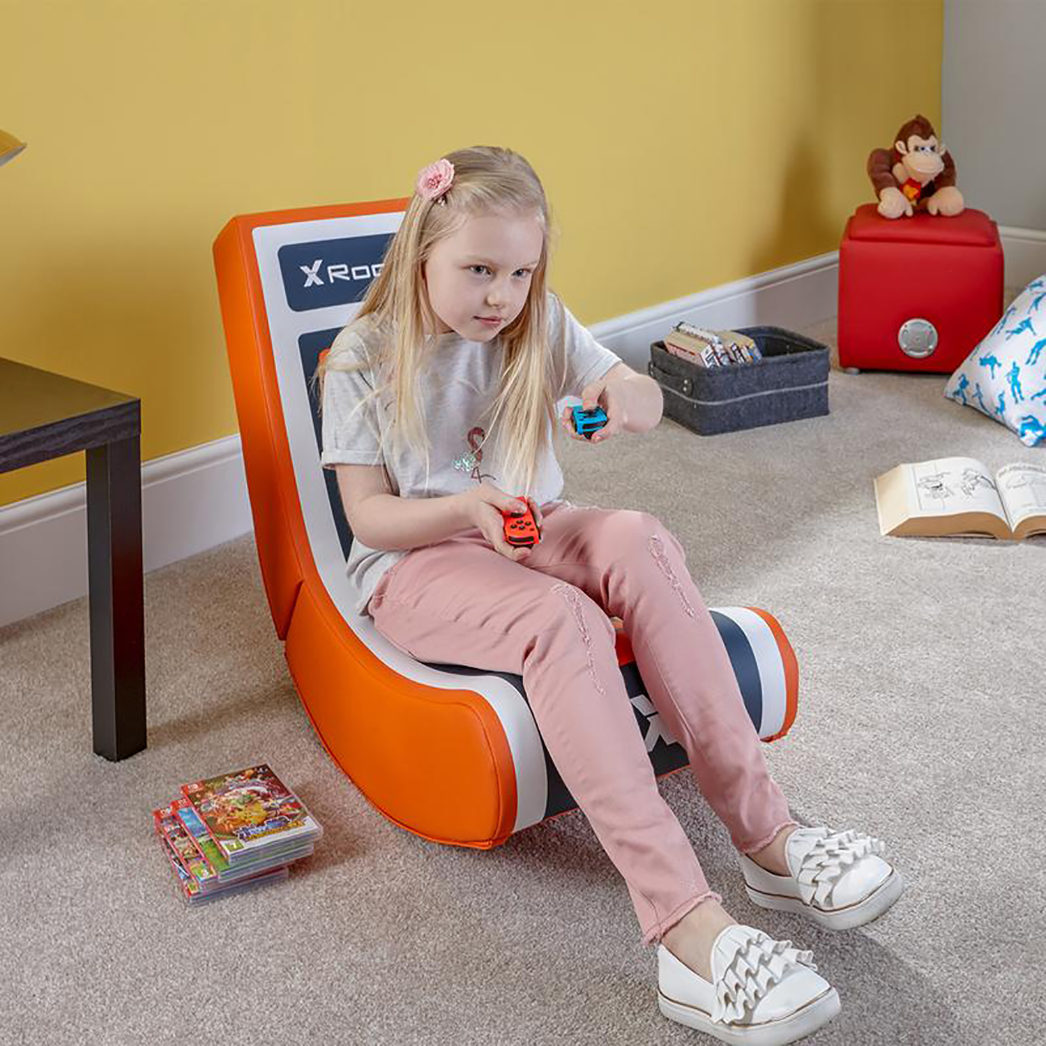 Ein junges Mädchen sitzt in einem orangefarbenen Gaming-Stuhl und hält zwei Spielzeugautos. Bücher und Spielzeug sind in der Nähe.