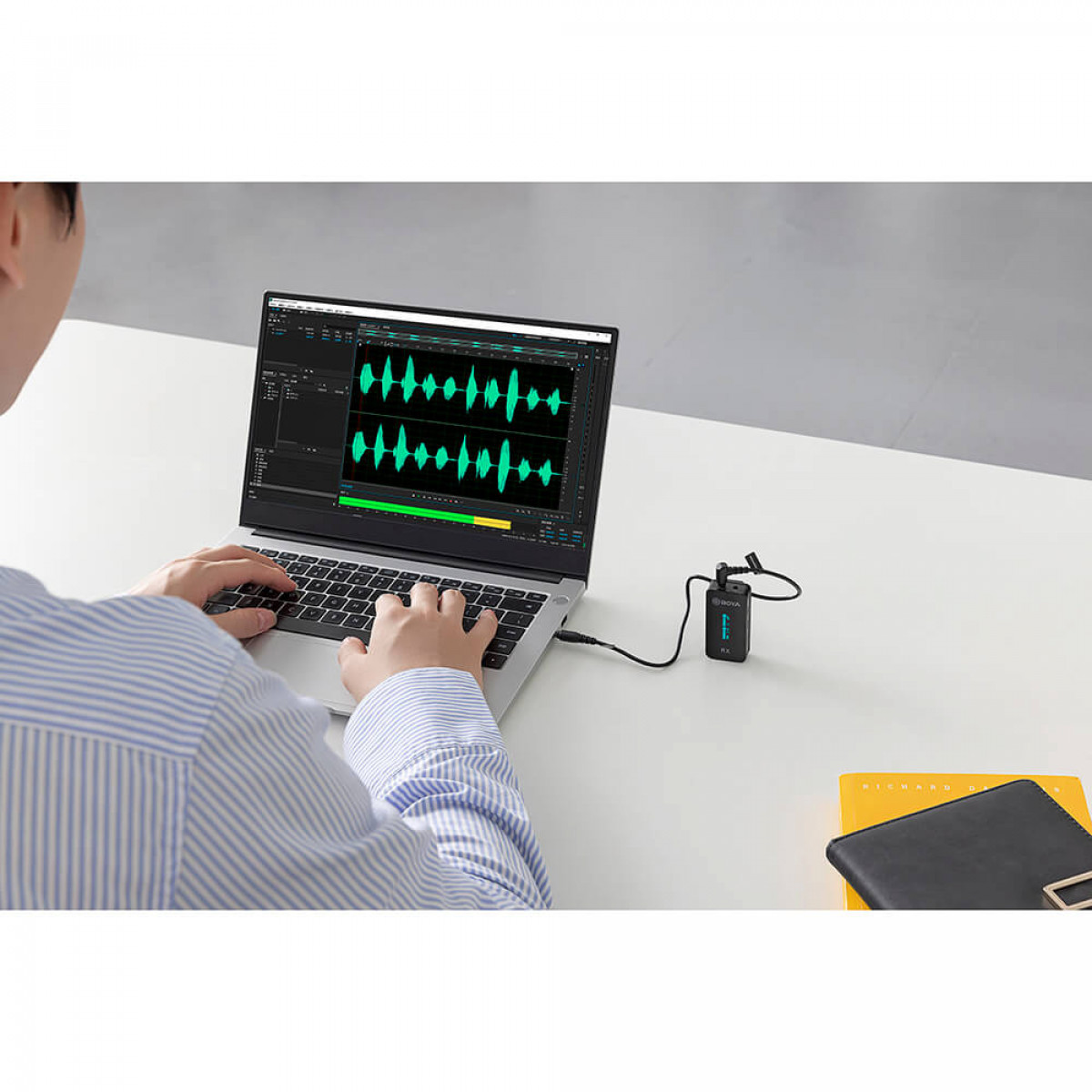 Person arbeitet an einem Laptop mit angeschlossenem Mikrofon. Der Laptop zeigt die Audio-Wellenform.