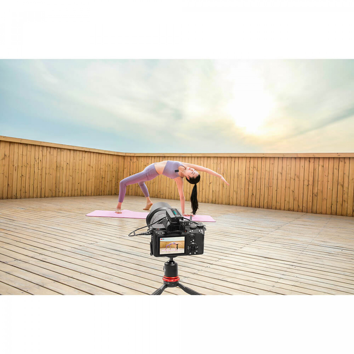Eine Frau macht Yoga auf einer rosafarbenen Matte auf einem Holzdeck. Eine Kamera ist eingerichtet, um sie aufzuzeichnen.
