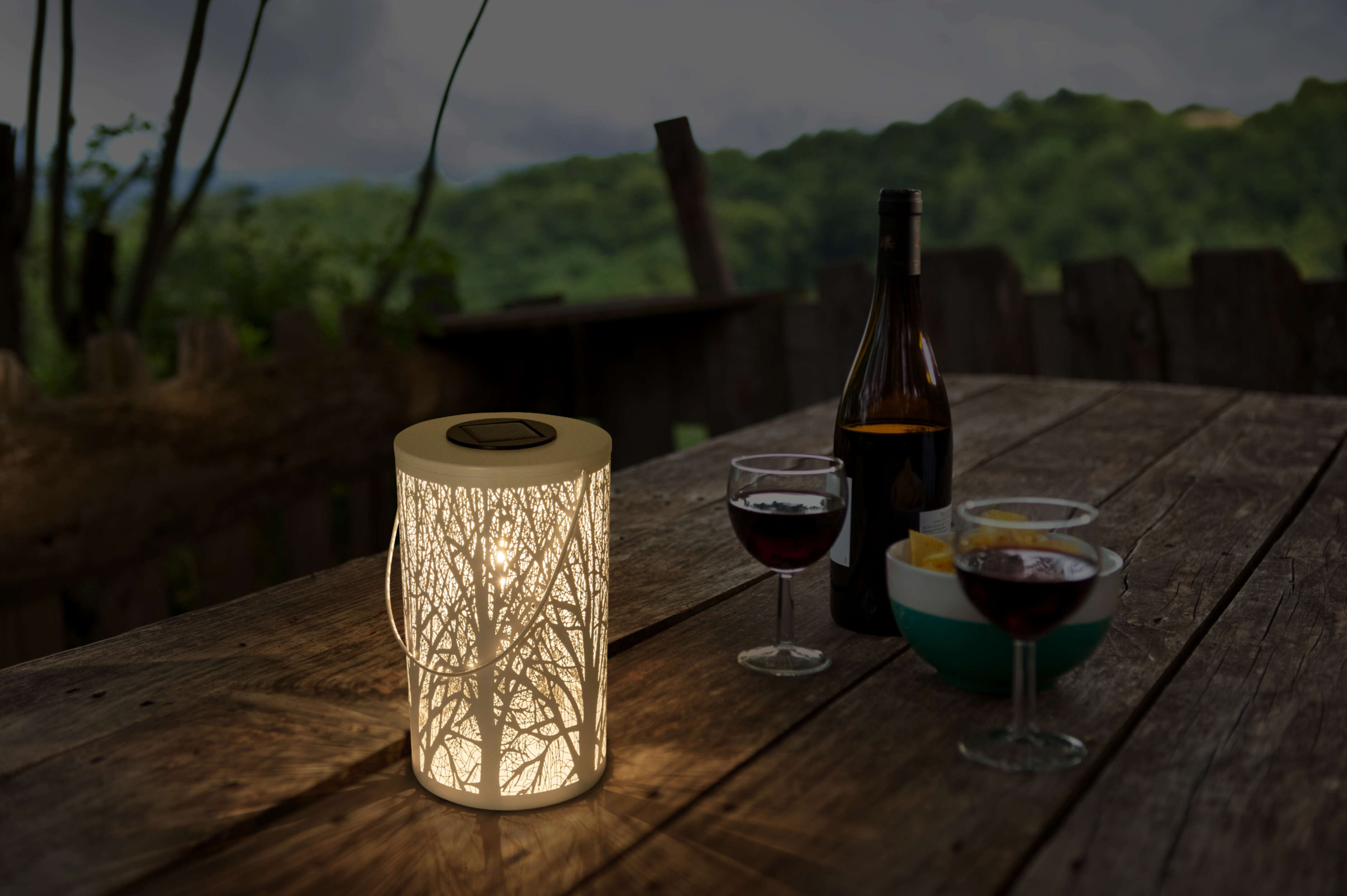 Laterne auf einem Tisch mit Wein, Flasche und Bäumen im Hintergrund.