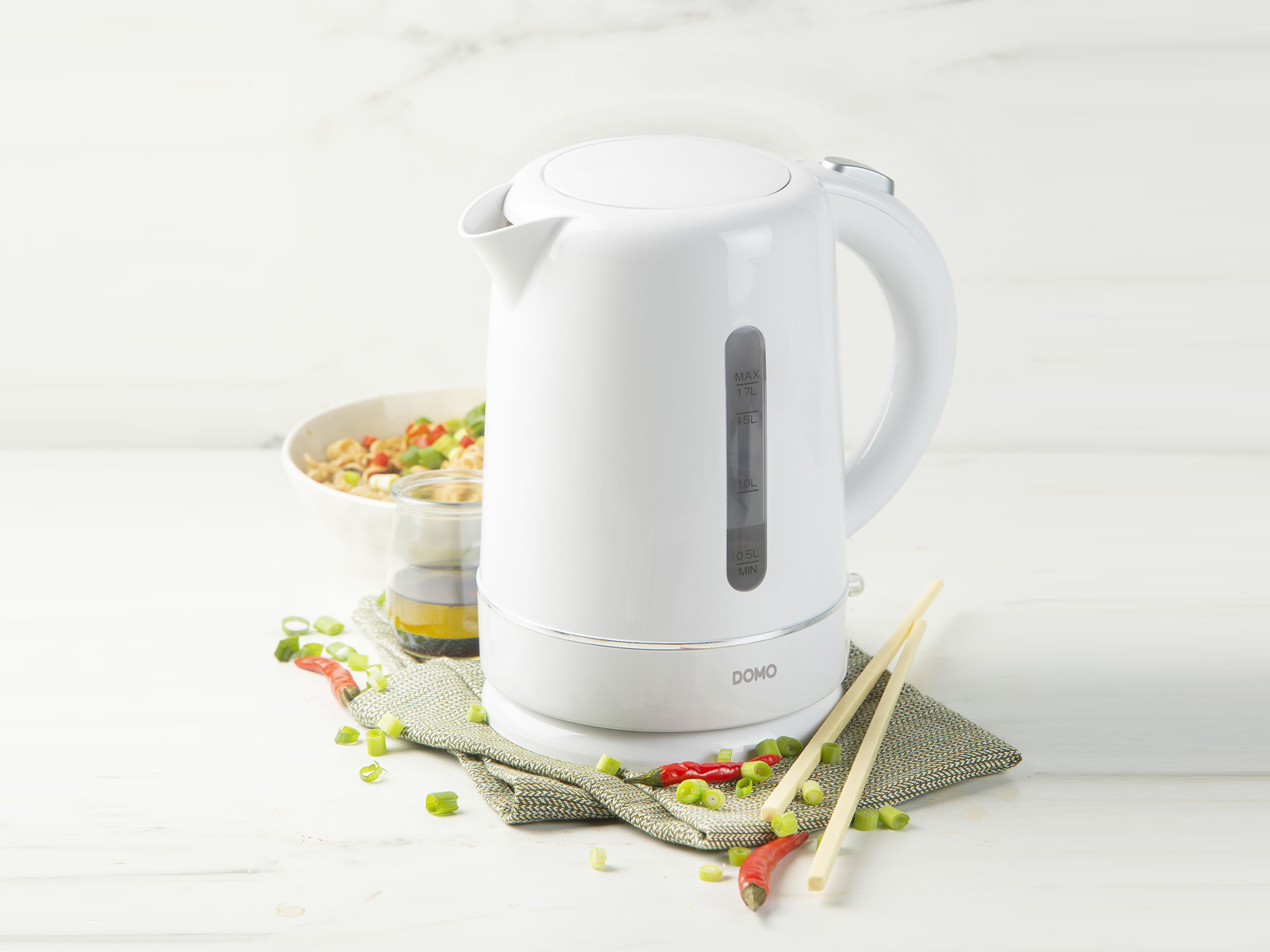 Biały czajnik elektryczny ze szklanką sosu, pałeczkami i miską jedzenia. Czerwone papryczki i szczypiorek.