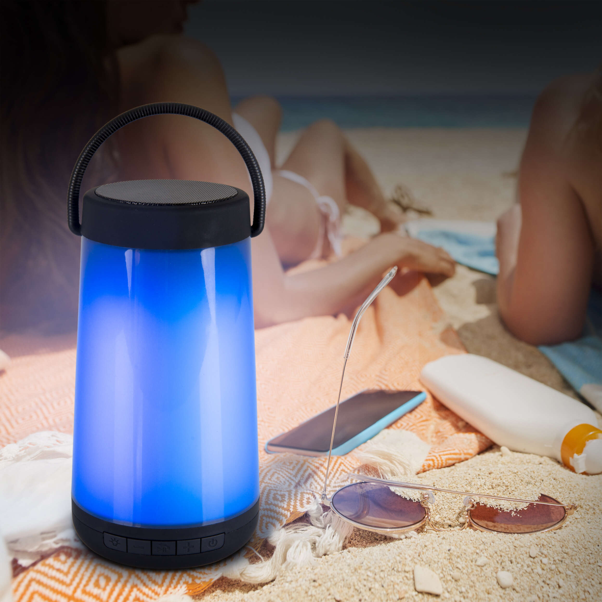 Blauer Leucht-Lautsprecher am Strand. Zwei Personen sonnen sich im Hintergrund. Sonnenbrille und Sonnencreme sind in der Nähe.