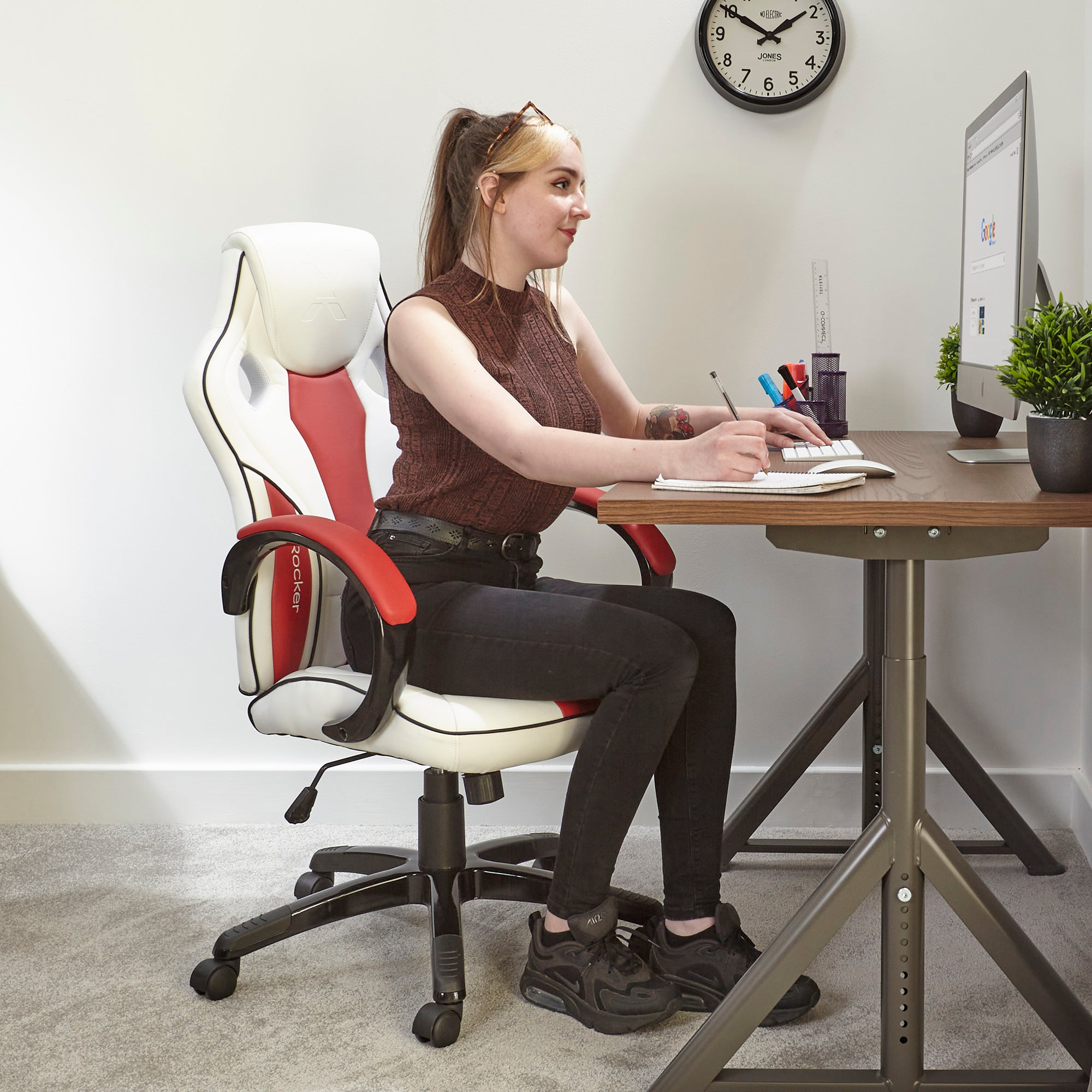 Frau sitzt an einem Schreibtisch vor einem Computer und schreibt in ein Notizbuch. Sie sitzt in einem Gaming-Stuhl.