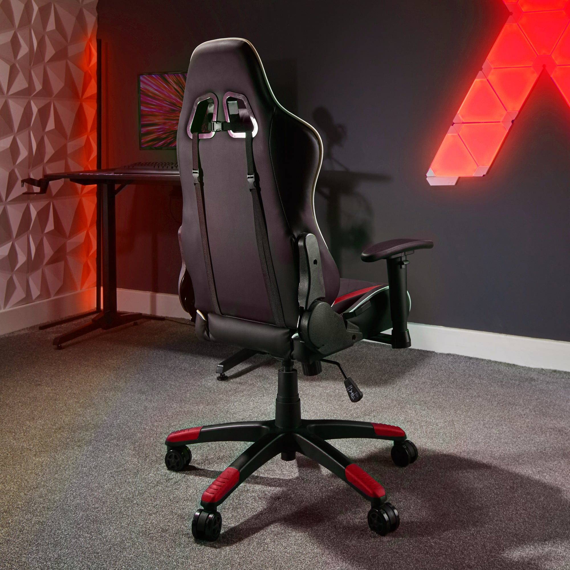 Ein Gaming-Stuhl von hinten. Er ist schwarz und rot. Der Stuhl steht auf einem grauen Teppich, mit Lichtern im Hintergrund.