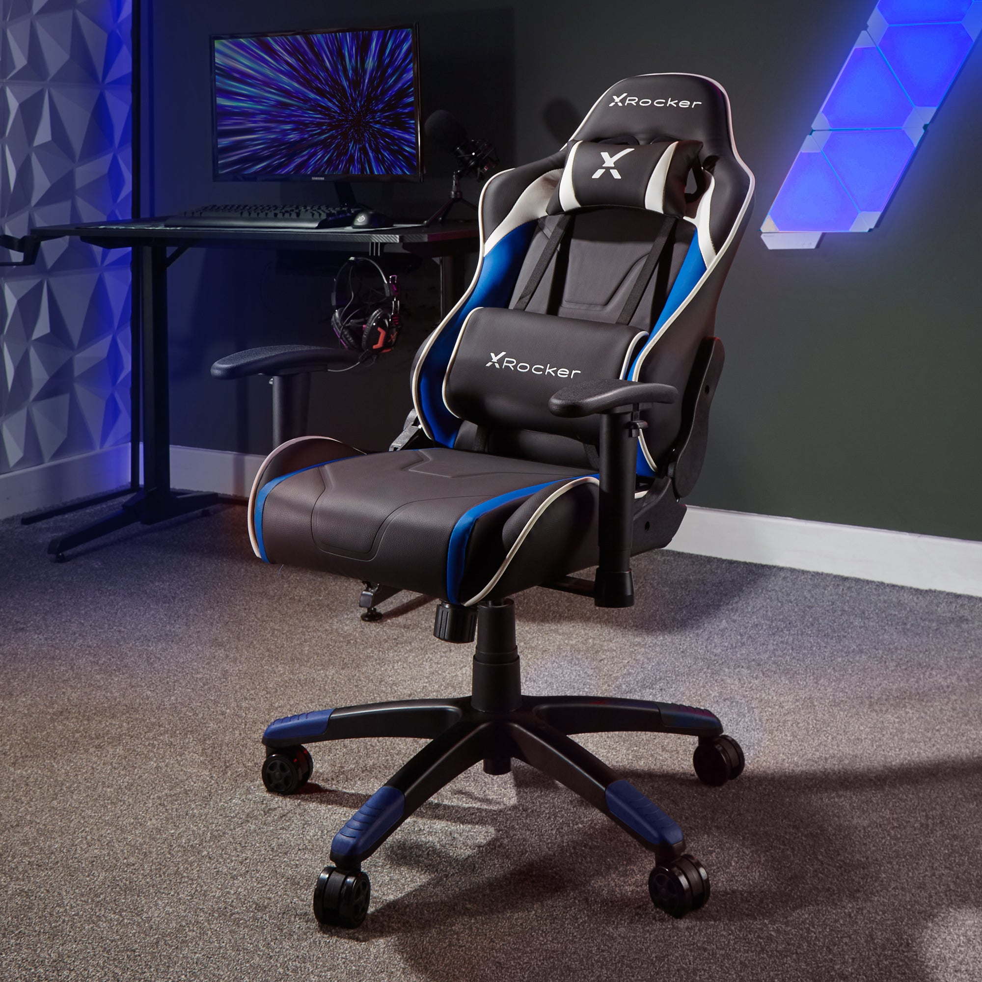 X Rocker Gaming-Stuhl. Schwarzer, blauer und weißer Stuhl auf grauem Teppich. Im Hintergrund befindet sich ein Computer-Setup.