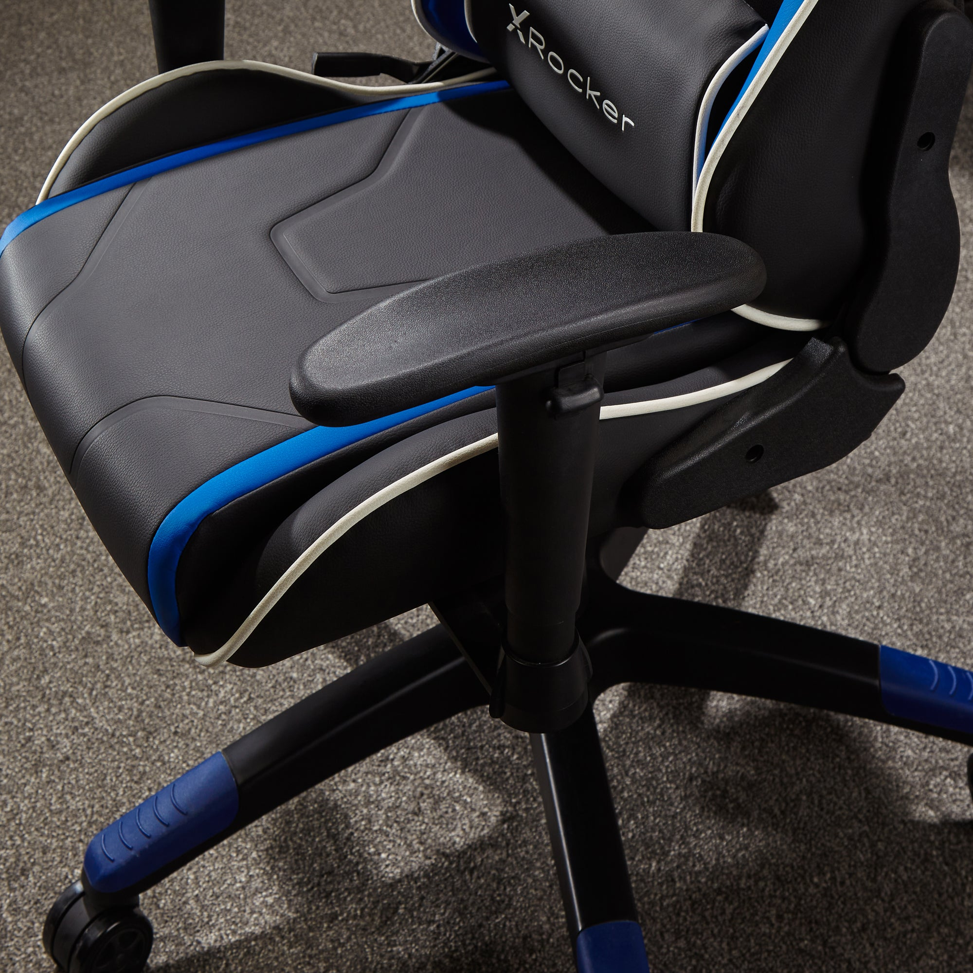 Ein schwarz-blauer Gaming-Stuhl mit X Rocker-Logo auf der Kopfstütze. Der Stuhl steht auf grauem Teppich.