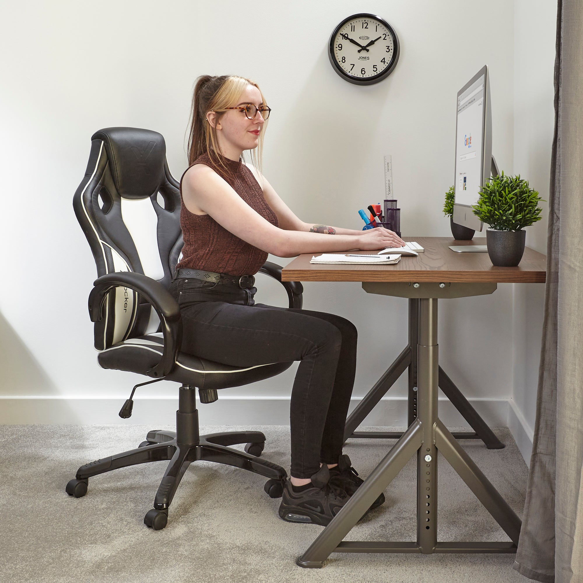 Frau am Schreibtisch, mit Brille, benutzt einen Computer. Eine Uhr und eine Pflanze sind an der Wand.