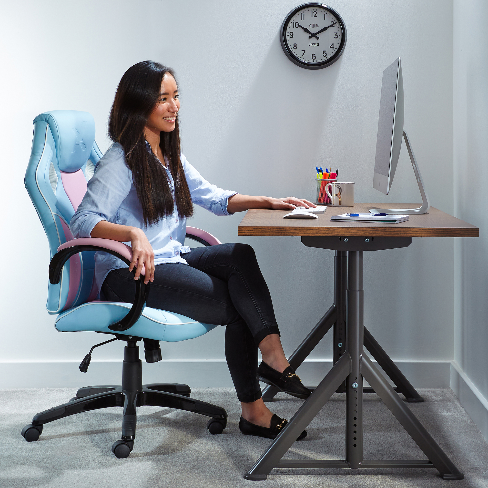 Frau in einem blau-rosa Gaming-Stuhl, arbeitet an einem Schreibtisch mit Computer und Uhr.