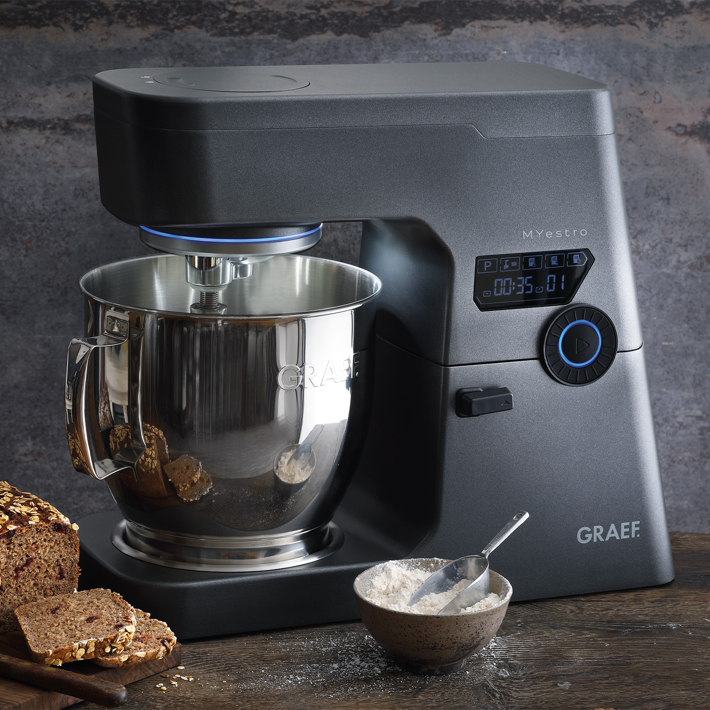 Ein grauer Küchenmixer mit einer Edelstahlschüssel, neben einem Brotlaib und einer Schüssel mit Mehl.