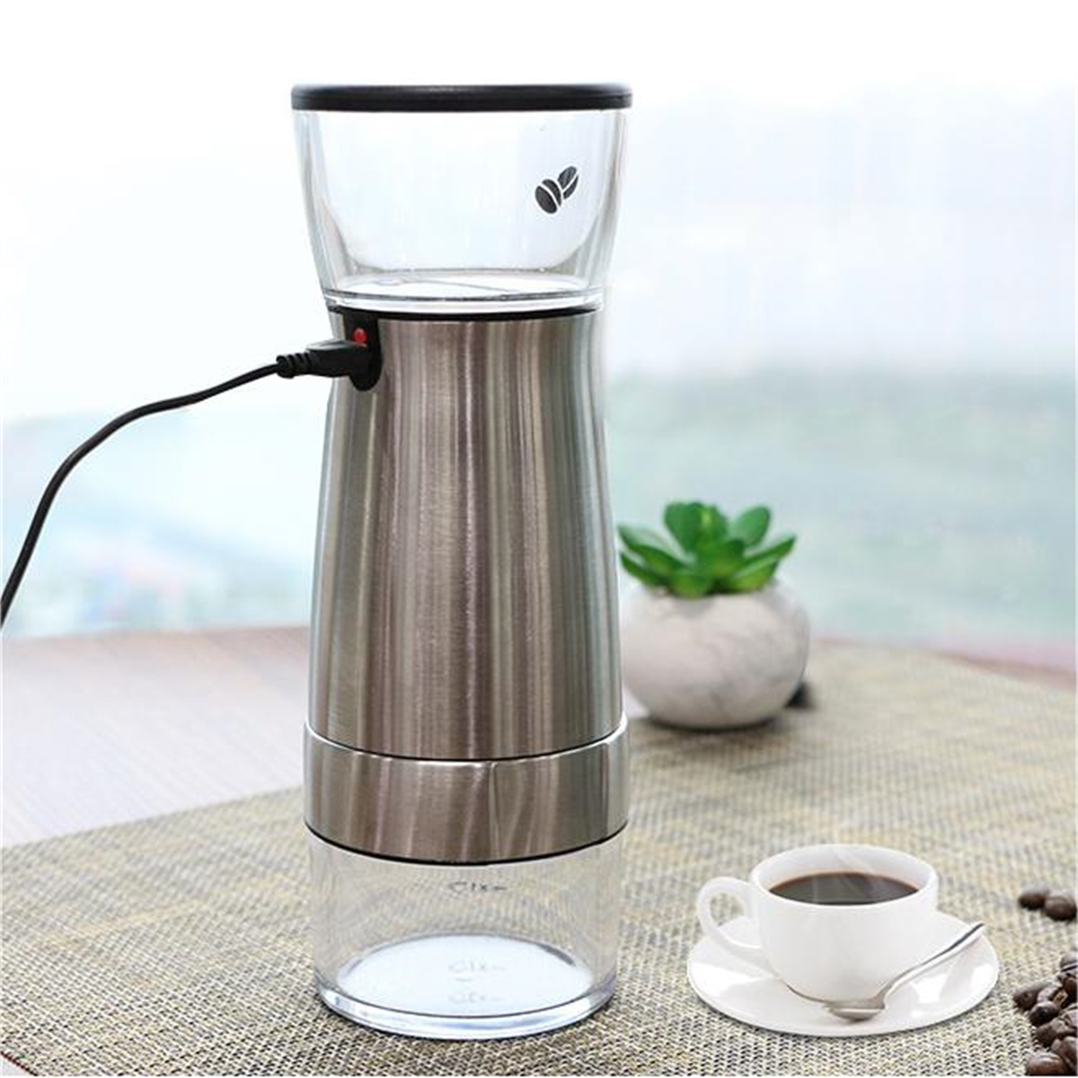 Eine Kaffeemühle aus Edelstahl mit klarem Ober- und Unterteil, Kaffeetasse und Pflanze.