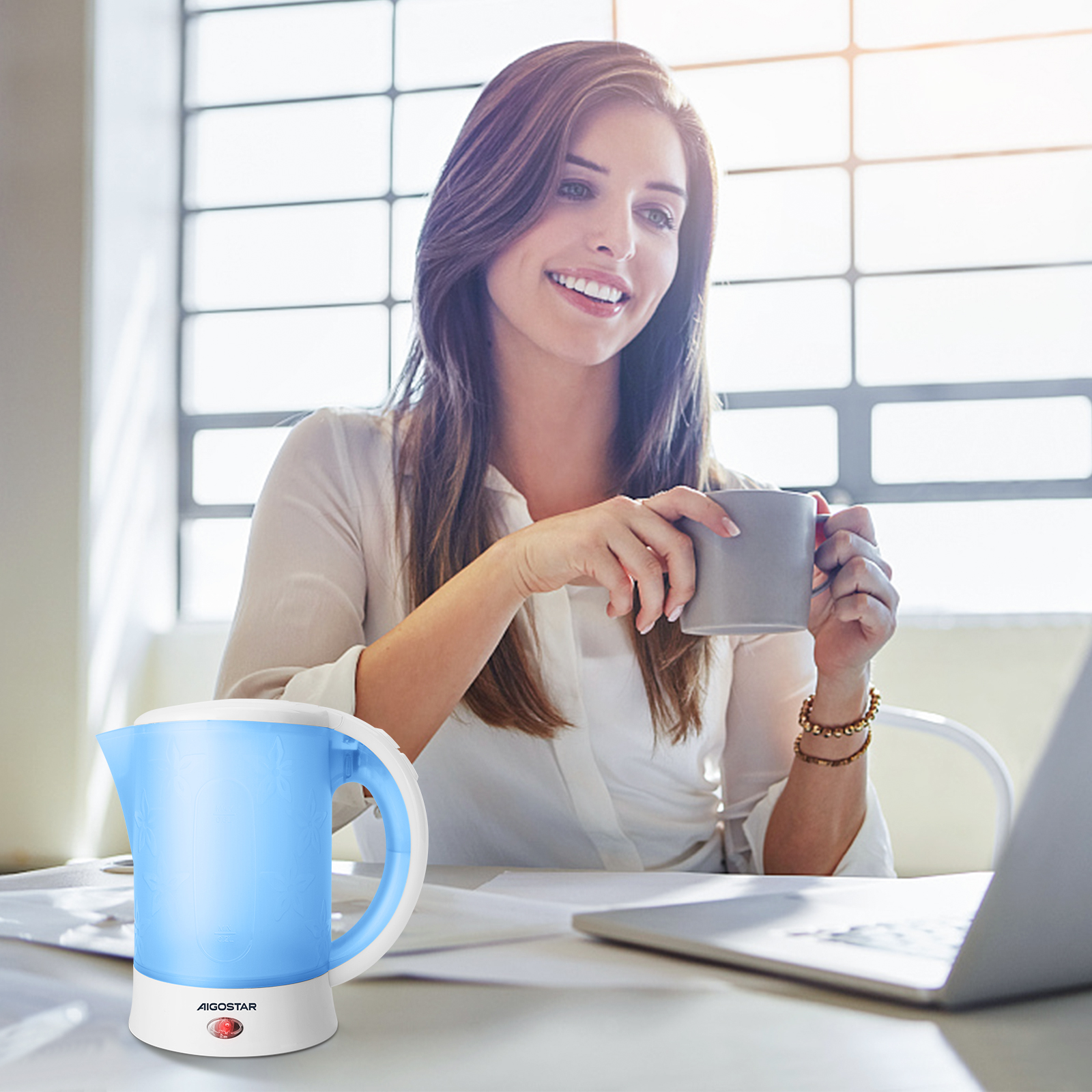 Frau lächelt, hält Tasse, schaut auf Laptop. Blau-weißer Wasserkocher auf dem Schreibtisch. Licht vom Fenster.