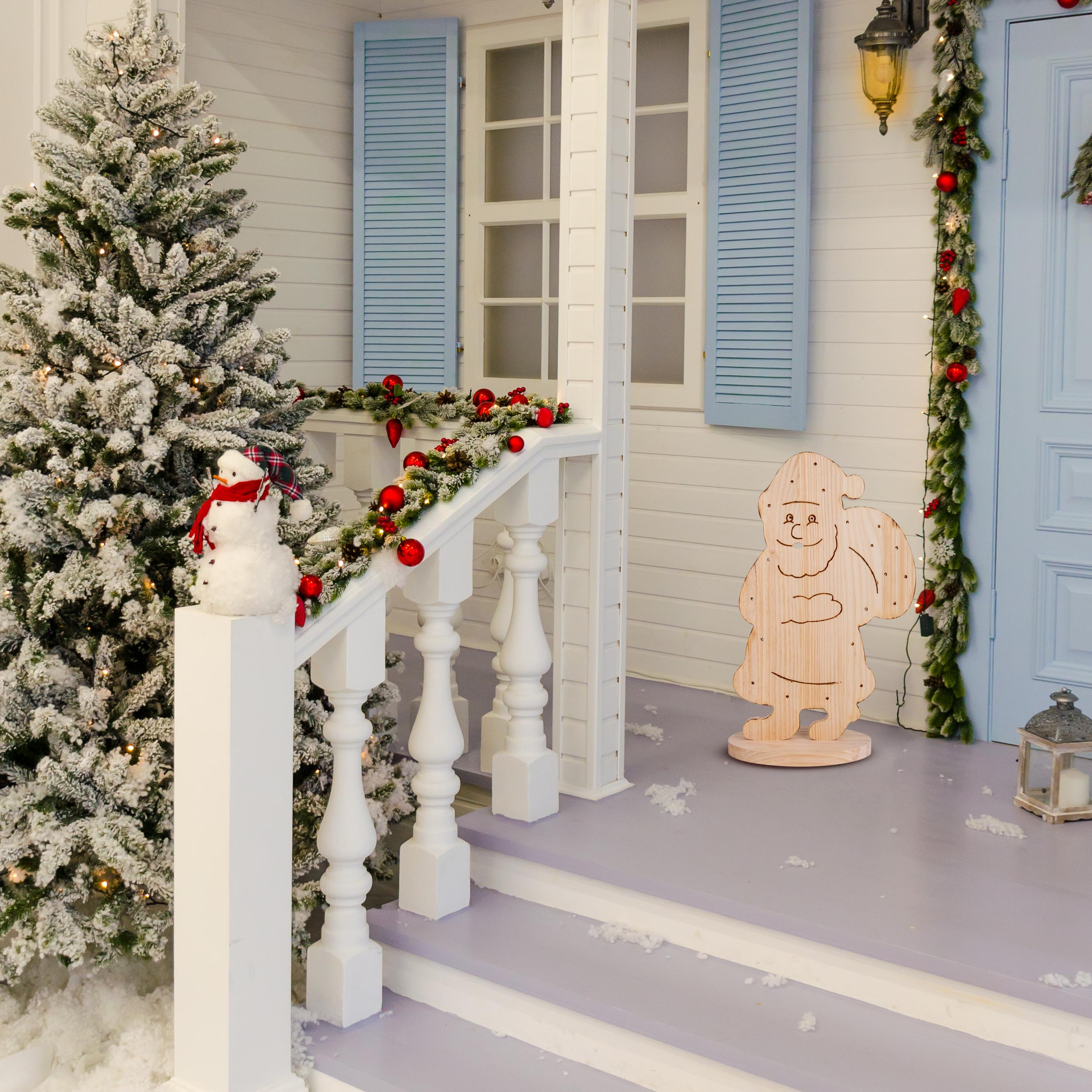 Eine verschneite Veranda, weihnachtlich geschmückt. Ein Schneemann, Baum und hölzerne Santa-Figur sind sichtbar.
