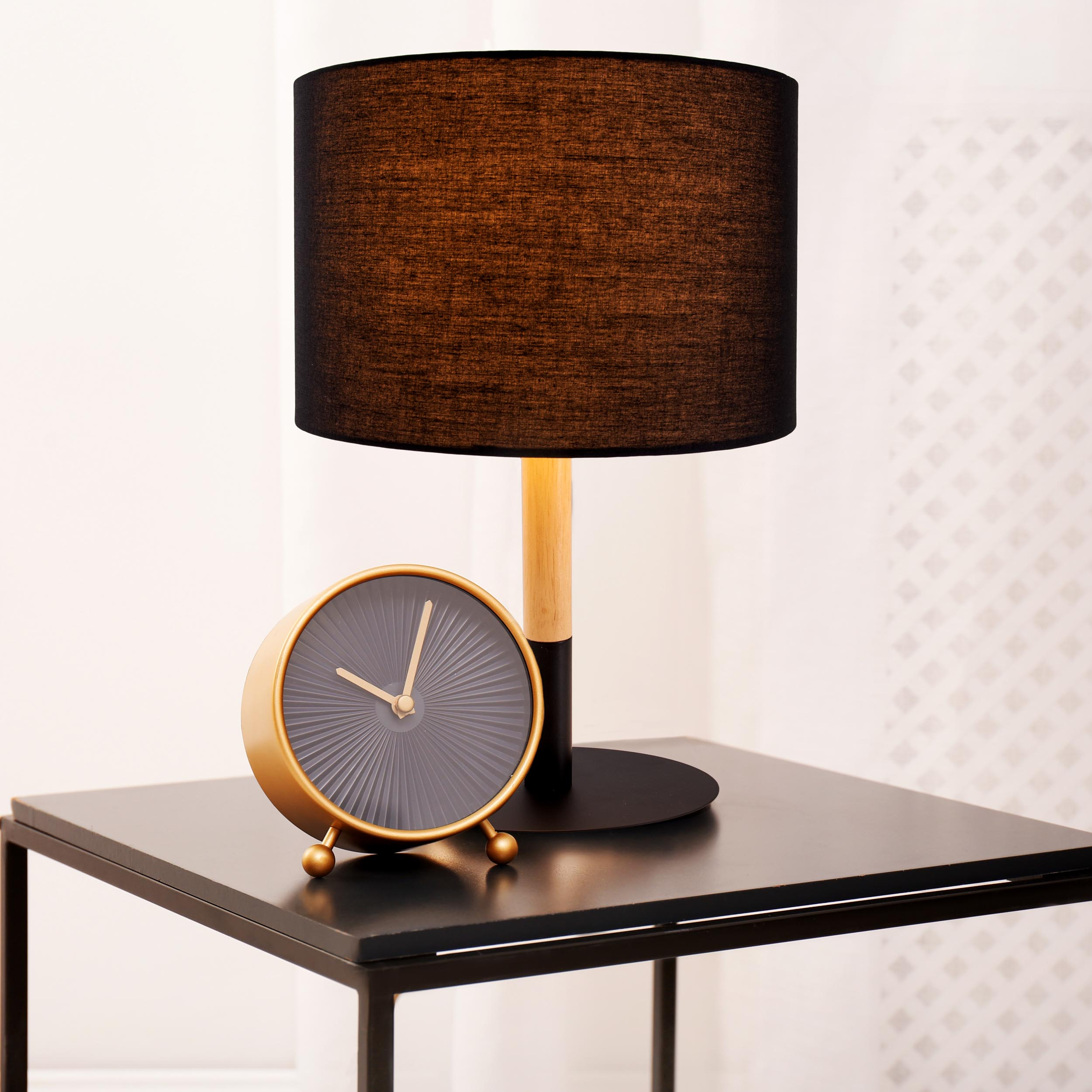 Tischlampe mit schwarzem Schirm auf einem schwarzen Tisch. Eine Uhr steht neben der Lampe.