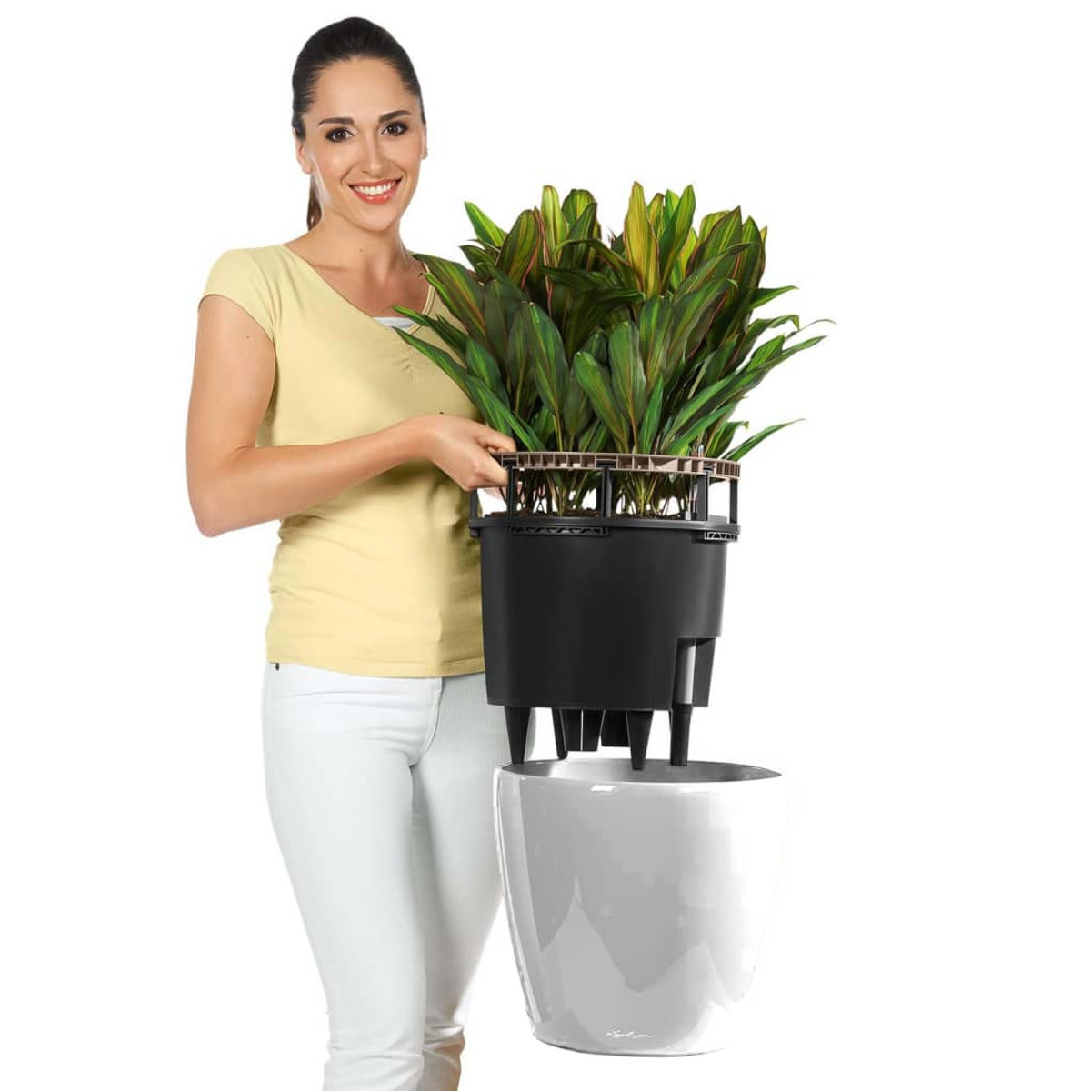 Femme tenant une plante dans un pot noir, placée dans un pot blanc, souriant à la caméra.