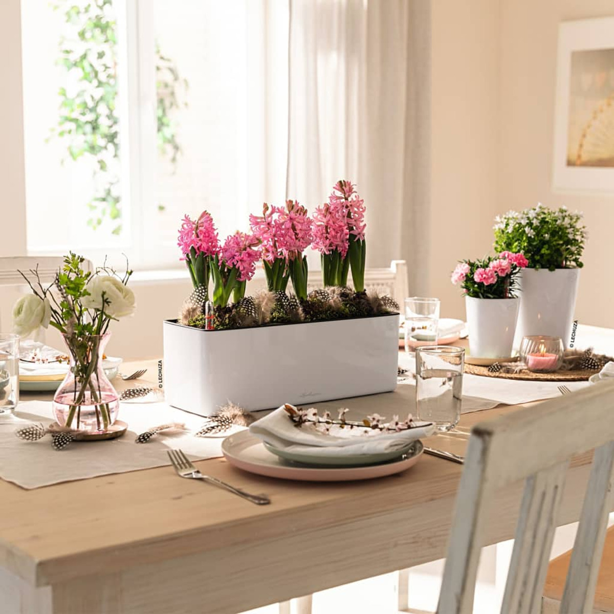 Table à manger dressée pour un repas, avec des fleurs roses et d'autres décorations. Une fenêtre en arrière-plan.