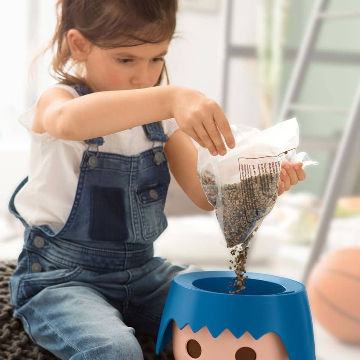 Enfant versant des graines dans un récipient bleu avec un visage. Un ballon de basket est à proximité.