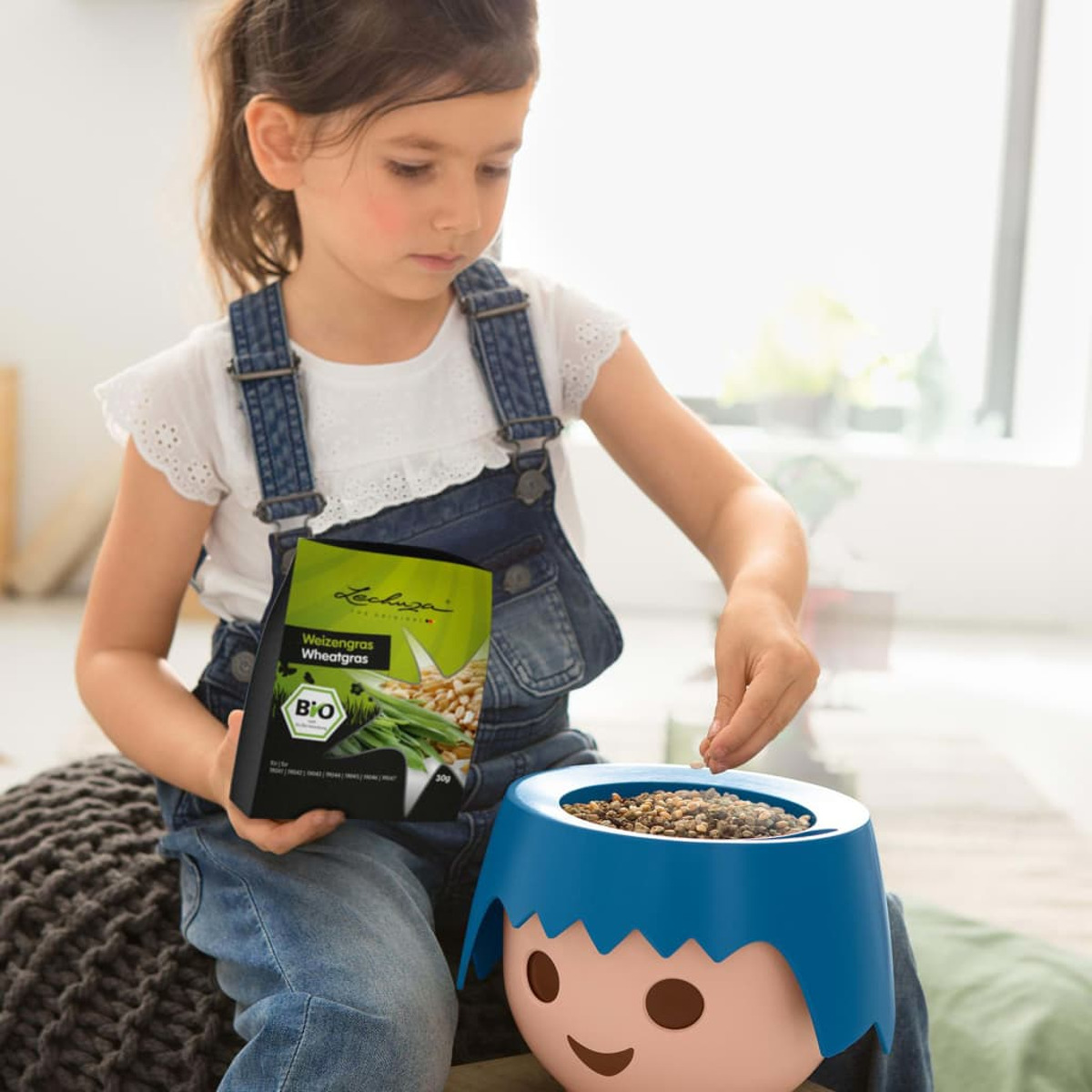 Une fille tient un paquet d'herbe de blé et met des graines dans un pot bleu.