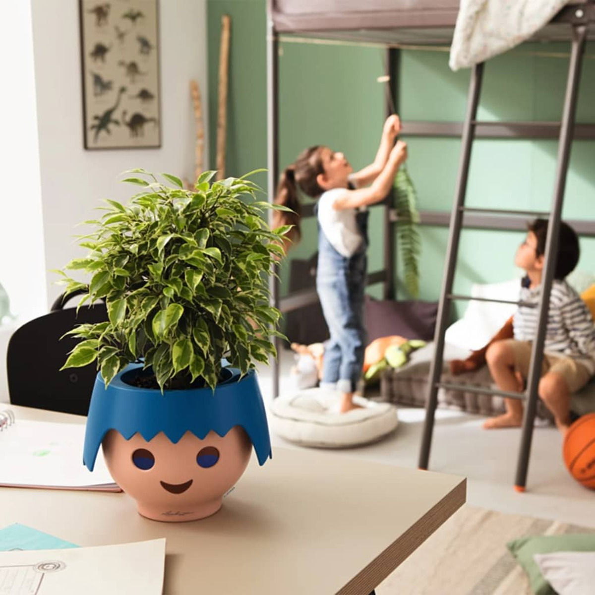 Une plante dans un pot en forme de visage sur un bureau. Des enfants jouent en arrière-plan.