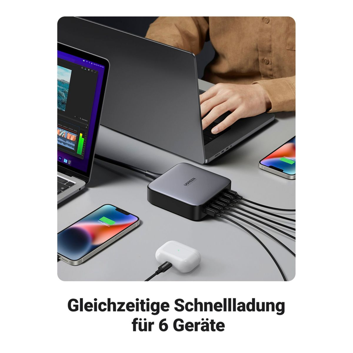 Grijze hub met kabels, twee laptops, twee telefoons opladen. Een persoon typt op een laptop.