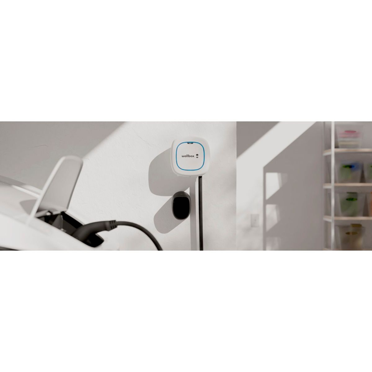 Weiß-blauer Wallbox-Lader an einer Wand montiert. Ein schwarzes Ladekabel hängt herunter.