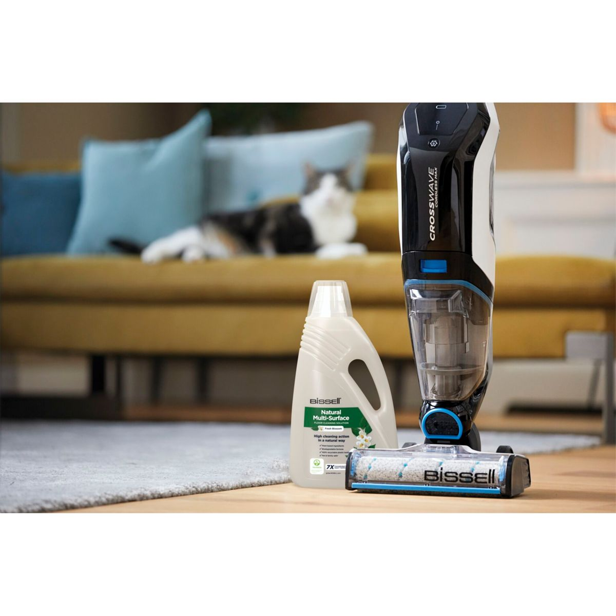 Aspirapolvere, liquido detergente e spazzola, con un gatto e un divano giallo sullo sfondo.