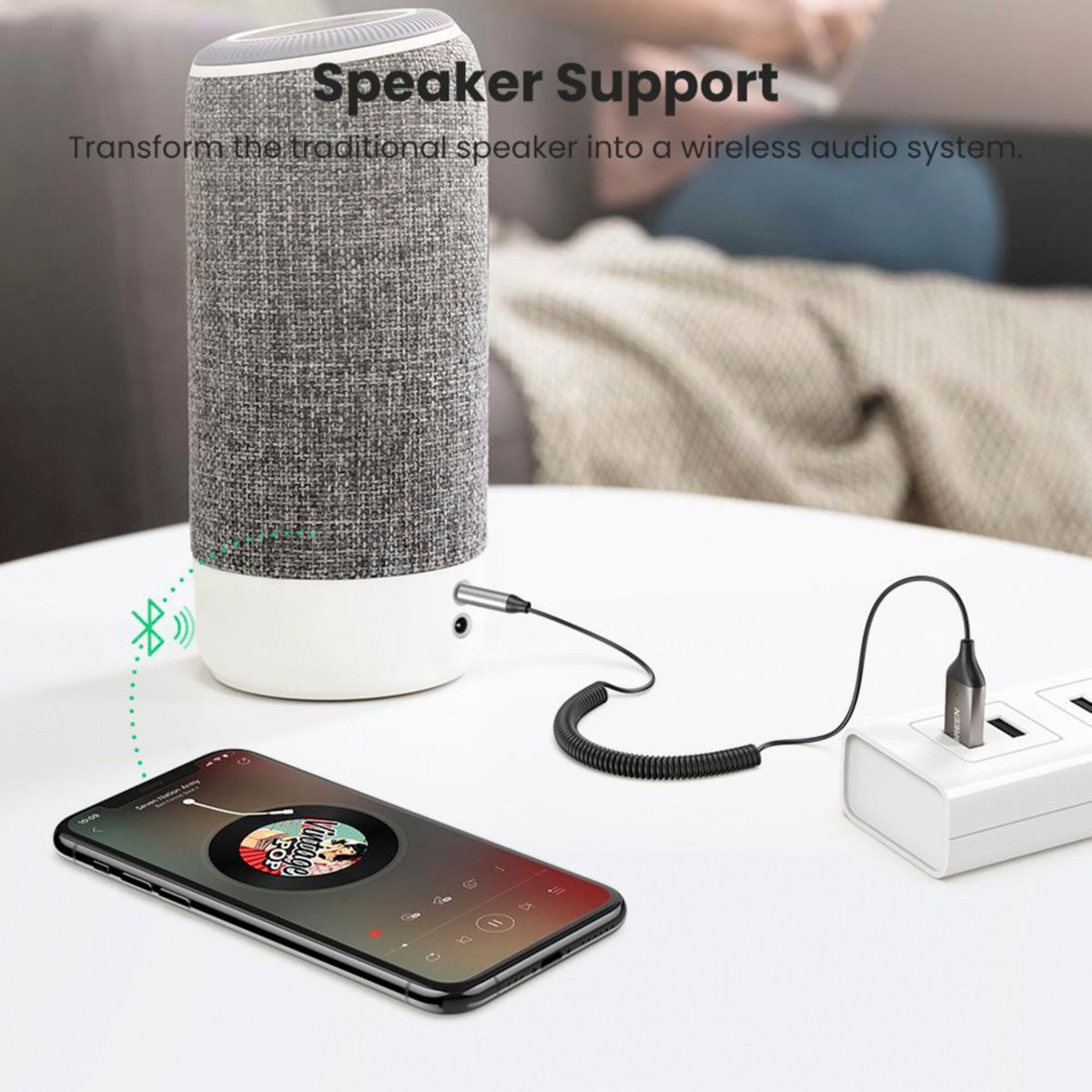 Bir telefonun ve bir güç adaptörünün yanında bir hoparlör. Telefon bir müzik uygulaması gösteriyor.