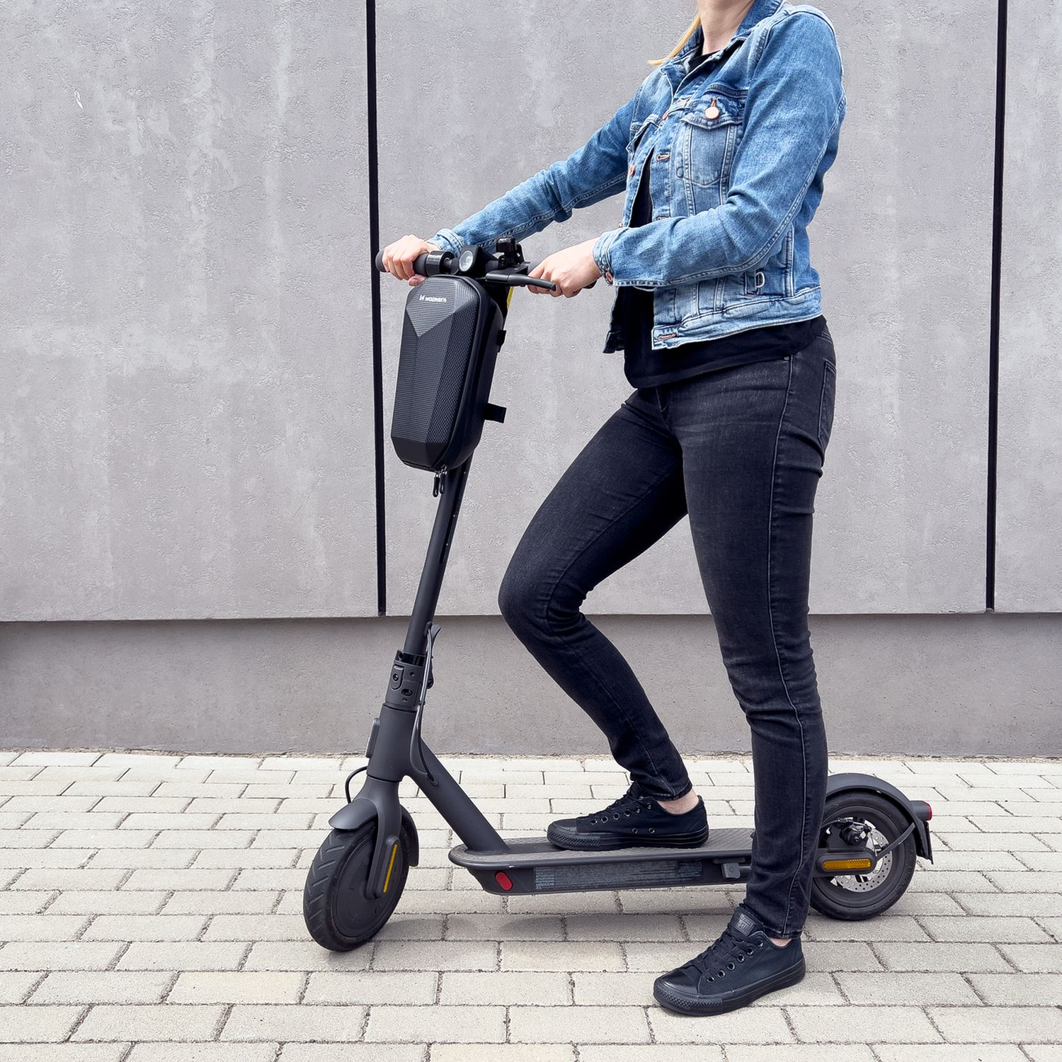 Una persona su un e-scooter, con una giacca di jeans e abiti neri, su un sentiero di mattoni.
