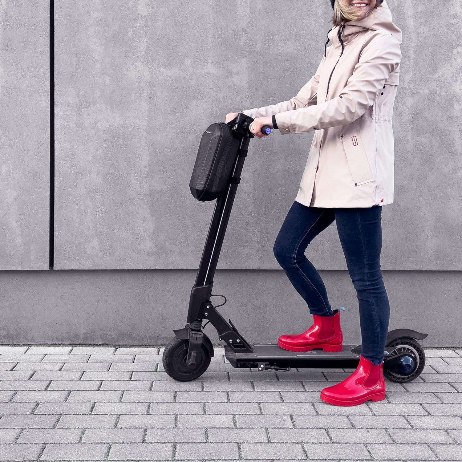 Donna su scooter con giacca e stivali rossi. Sorride, sfondo muro grigio.